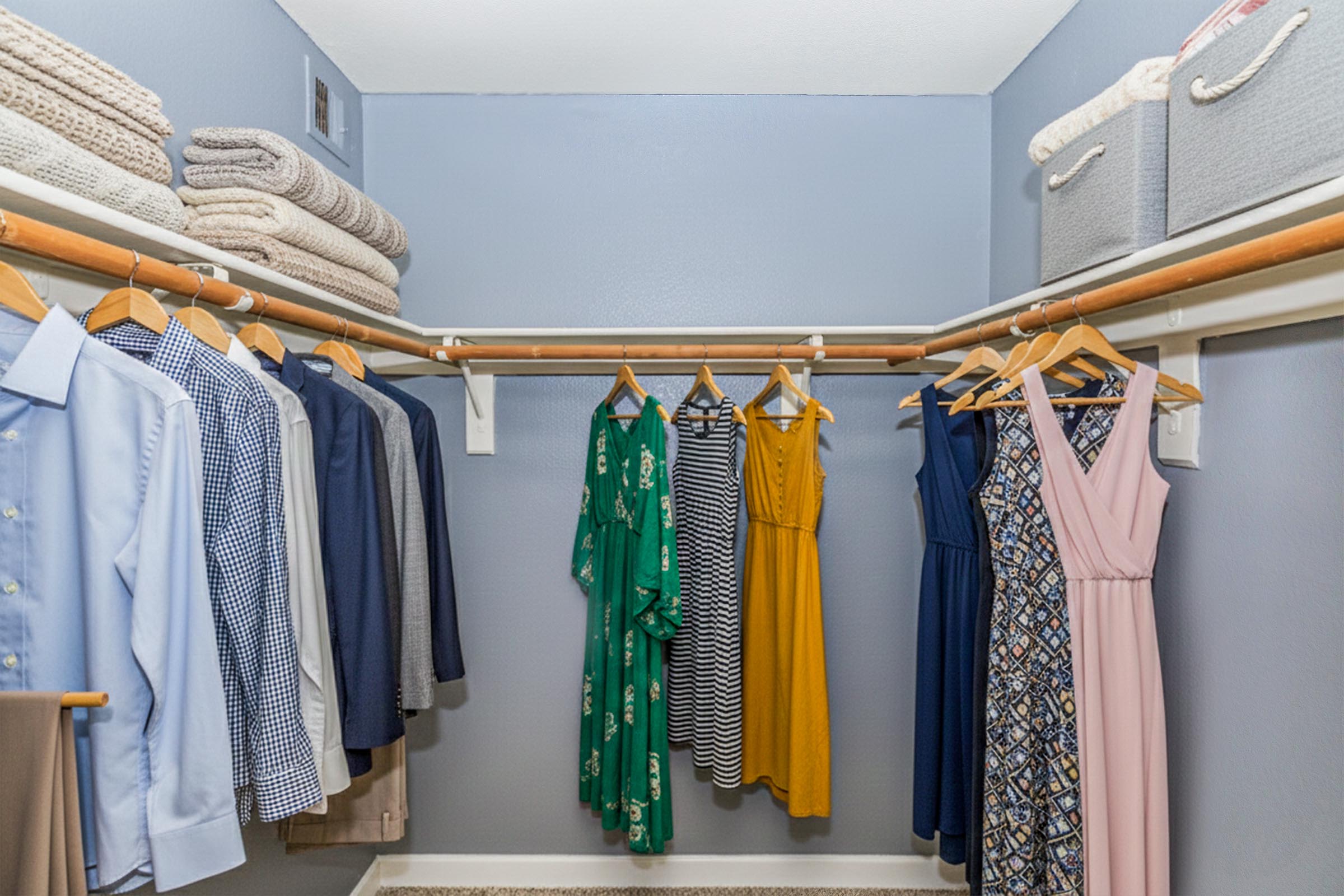 A neatly organized closet featuring a variety of clothing. On the left side, there are button-up shirts and blouses hanging, while the right displays several dresses in different colors and patterns. Towels are stacked on top shelves, and storage bins are neatly placed at the top. The walls are painted a soft blue.