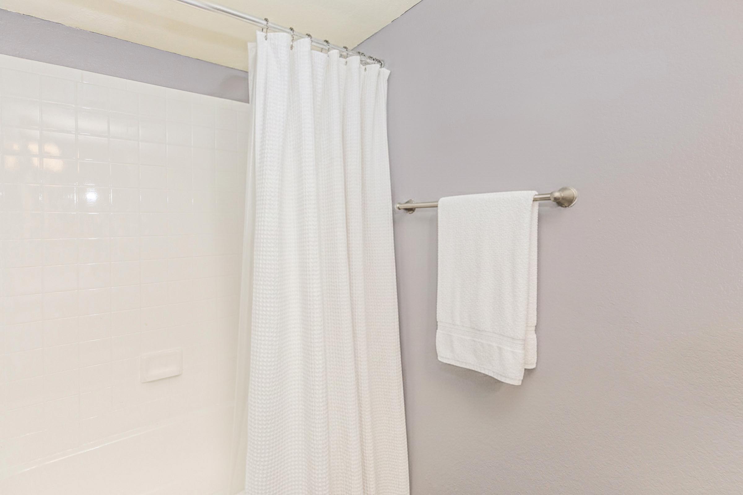 A bathroom scene featuring a white shower curtain hanging in front of a bathtub. A white towel is neatly hanging on a towel bar mounted on the wall. The wall is painted light gray, and the overall atmosphere is clean and simple.