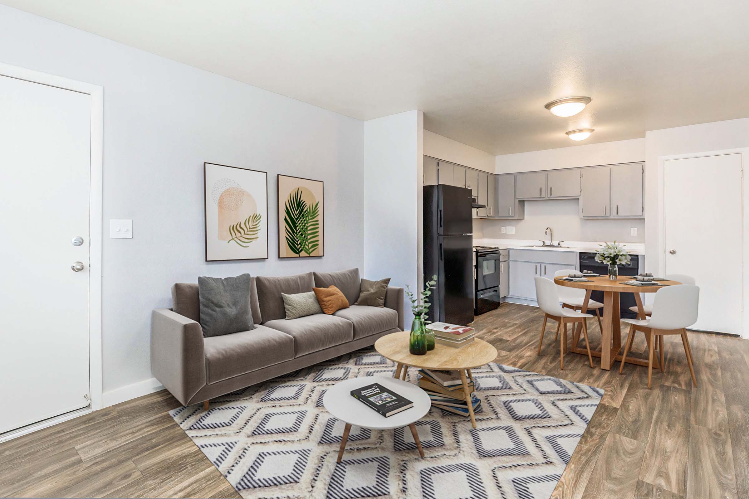 A modern living room and kitchen area featuring a gray sofa with decorative pillows, a round wooden coffee table, and a dining table with white chairs. The walls are light gray, and there are two framed wall art pieces. A door leads to another room, and the kitchen area has gray cabinets and black appliances.