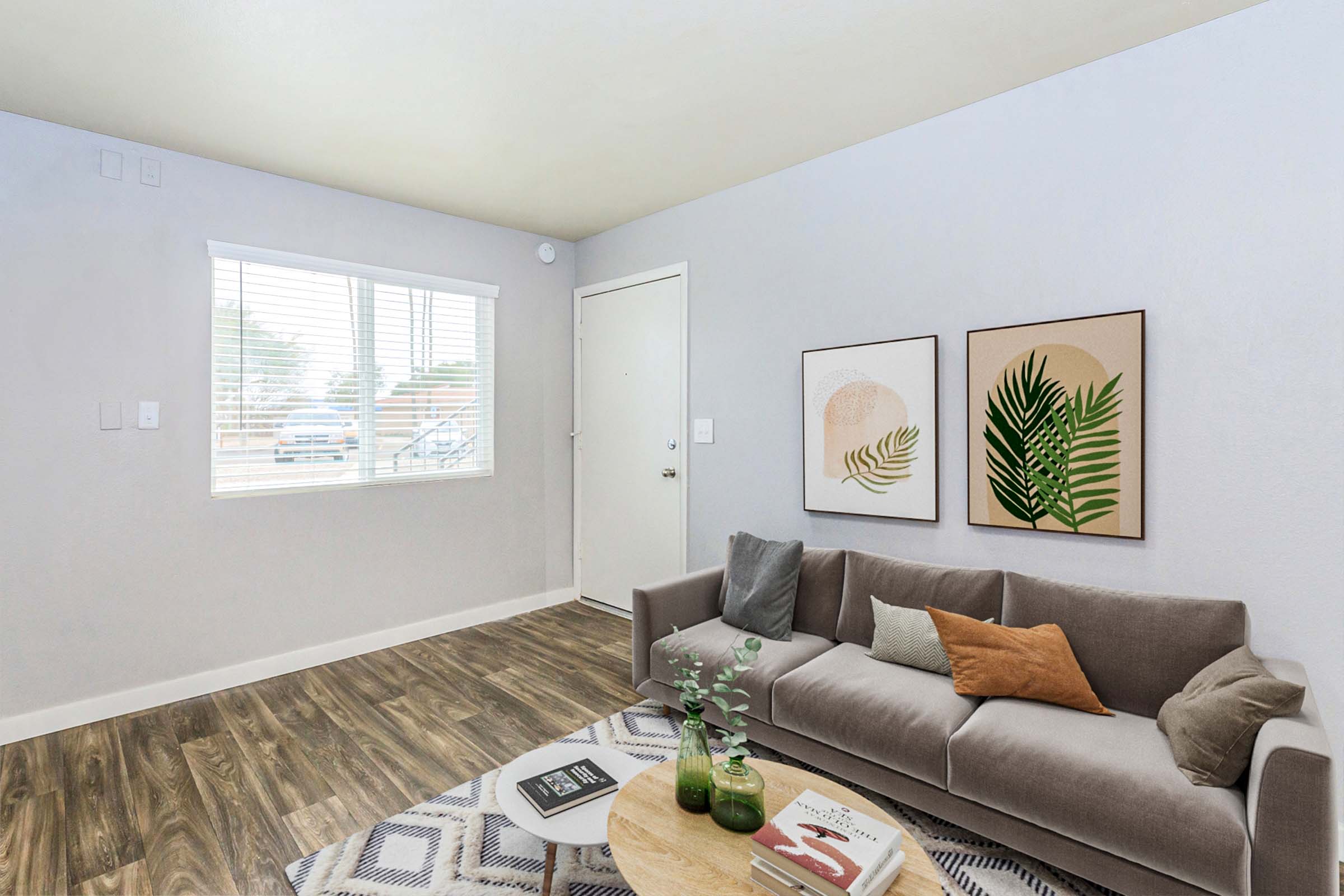 A cozy living room featuring a gray sofa with throw pillows in earthy tones, a round coffee table with books and a plant, and two framed art pieces on the wall. Natural light floods in through a window with white blinds, illuminating the room's modern decor and warm wood-like flooring.