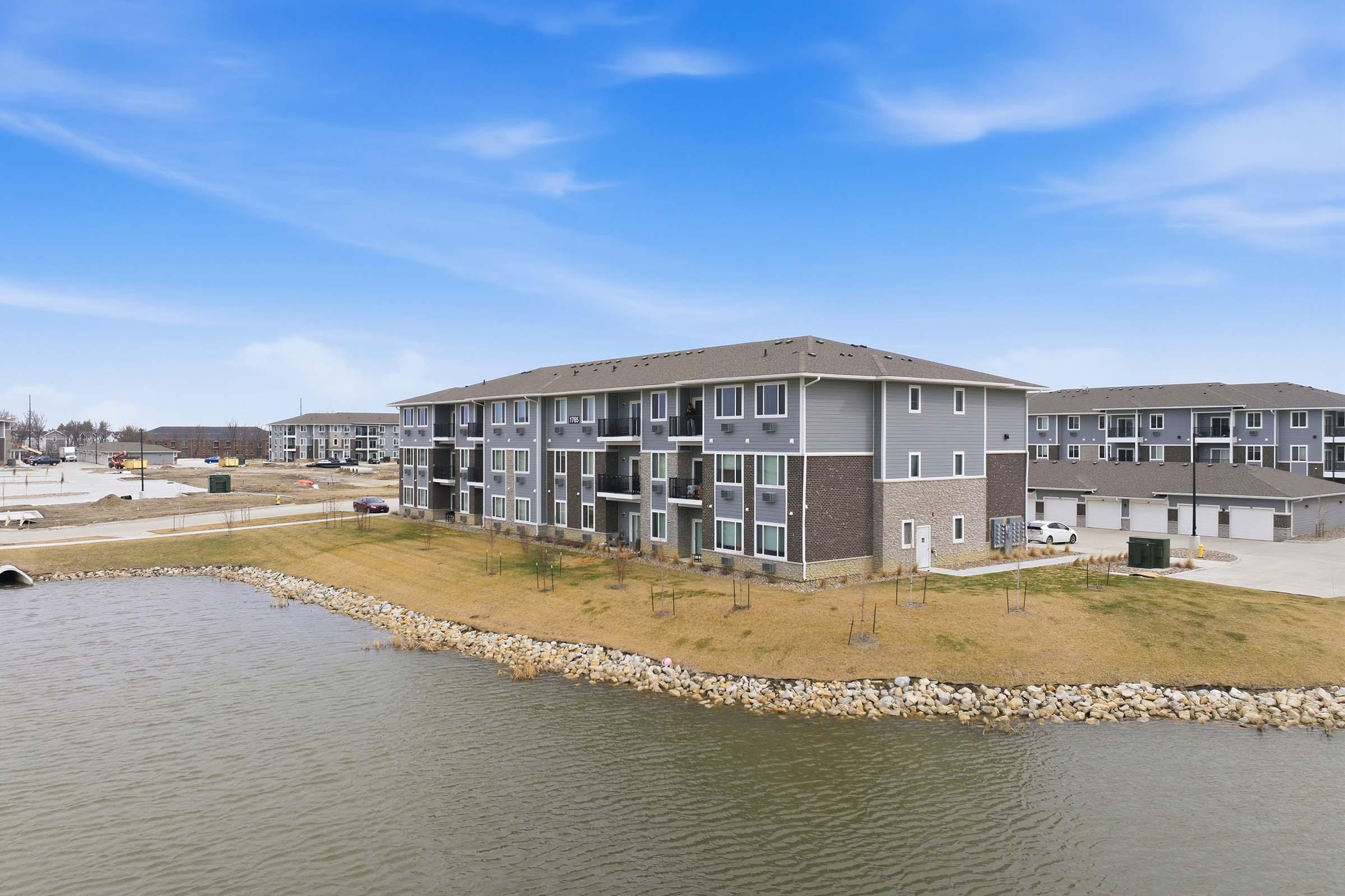 A modern multi-unit apartment building located by a calm water body, surrounded by green grass and rocky shoreline. The sky is bright blue with a few clouds, and additional residential buildings are visible in the background.
