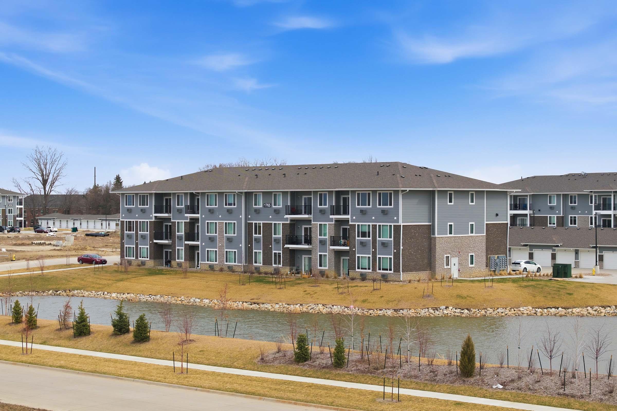A modern multi-unit apartment complex beside a small pond, featuring a landscaped area with grass and young trees. The building has a mix of gray and stone exterior with balconies. Clear blue skies above create a bright and inviting atmosphere.