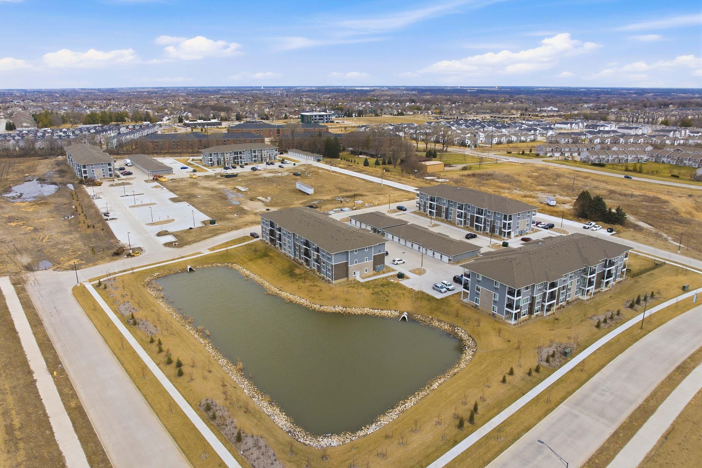 Aerial view of a residential area featuring multiple apartment buildings arranged around a small pond. The landscape includes parking lots, grassy patches, and nearby streets. In the background, additional housing and open space are visible under a clear blue sky.