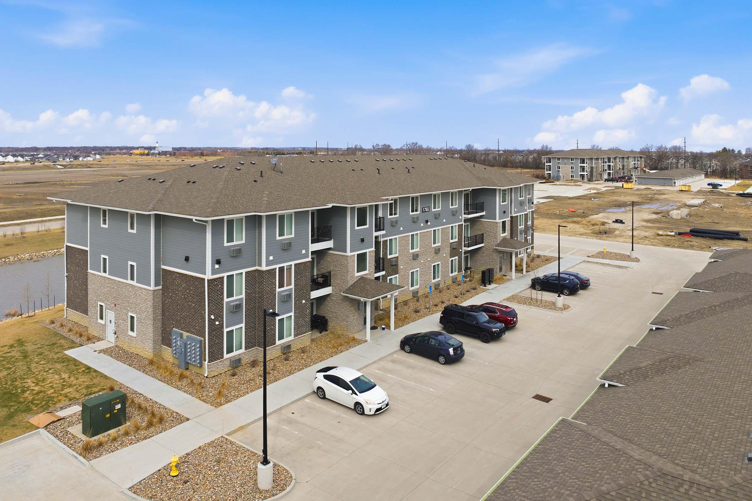 Aerial view of a multi-unit residential building with a mix of brick and siding, surrounded by a parking lot with several cars. In the background, additional buildings and an undeveloped area with construction materials are visible under a clear blue sky.