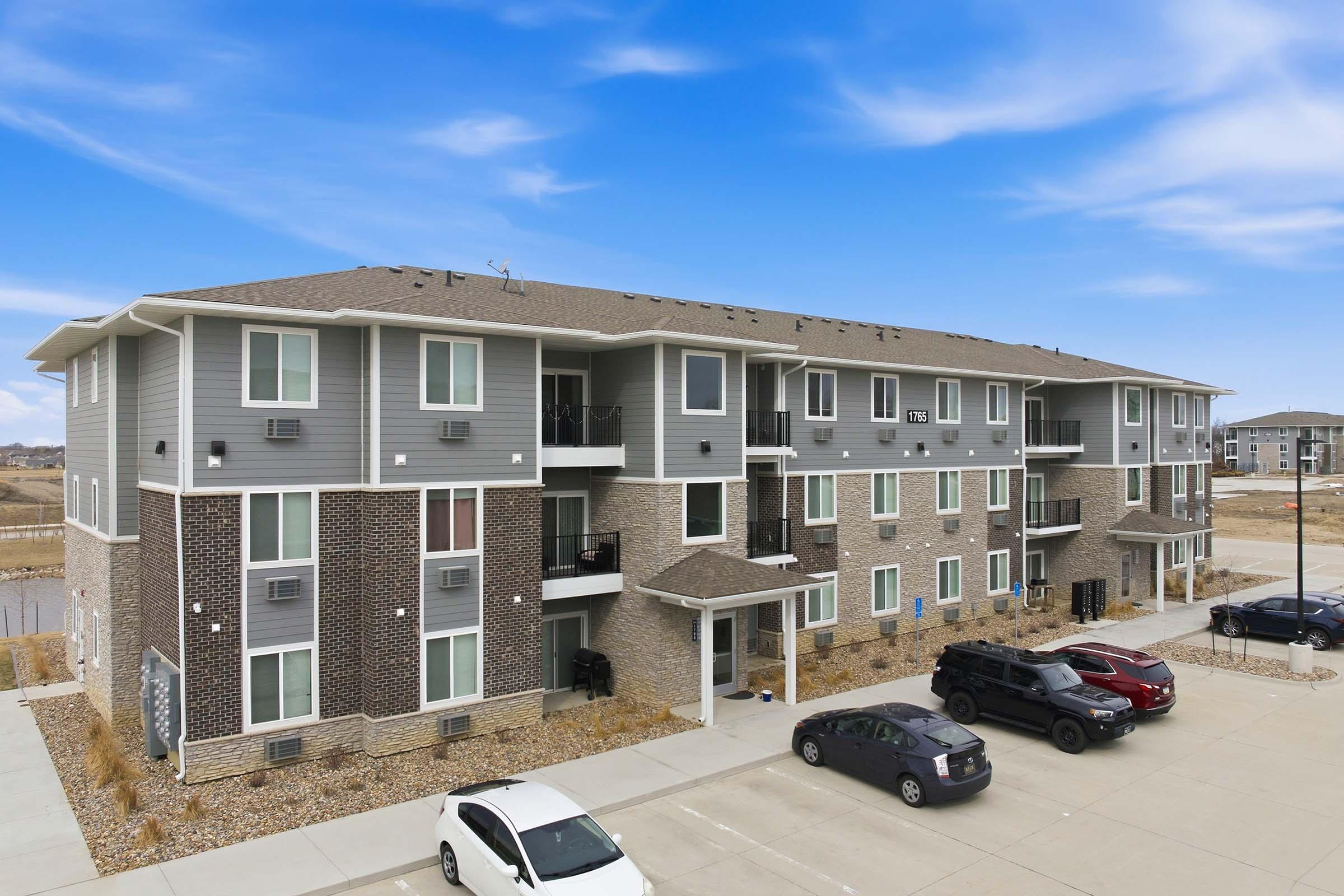 A three-story apartment building featuring a combination of brick and siding exteriors. The building has balconies and several windows. In the foreground, there is a parking lot with a few cars parked. The sky is clear with a blue background, and the surroundings are mostly open land.