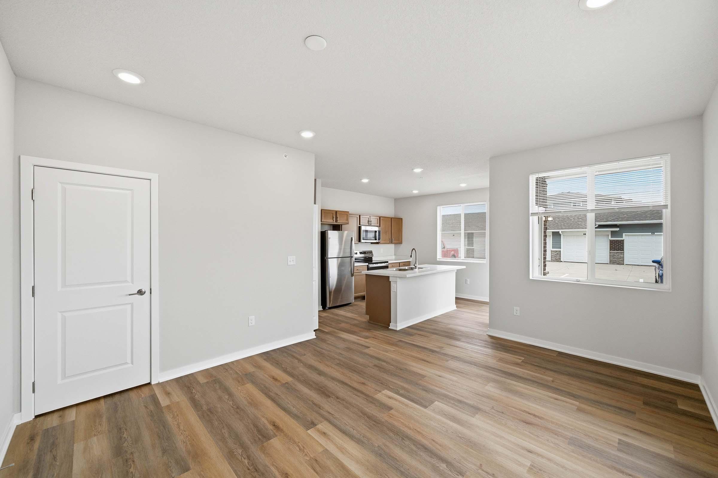 A spacious, modern living area featuring light-colored walls, recessed lighting, and a wood-style floor. On the left, there's a door leading to another room, and in the background, a kitchen with stainless steel appliances and an island. Large windows let in natural light, creating a bright atmosphere.
