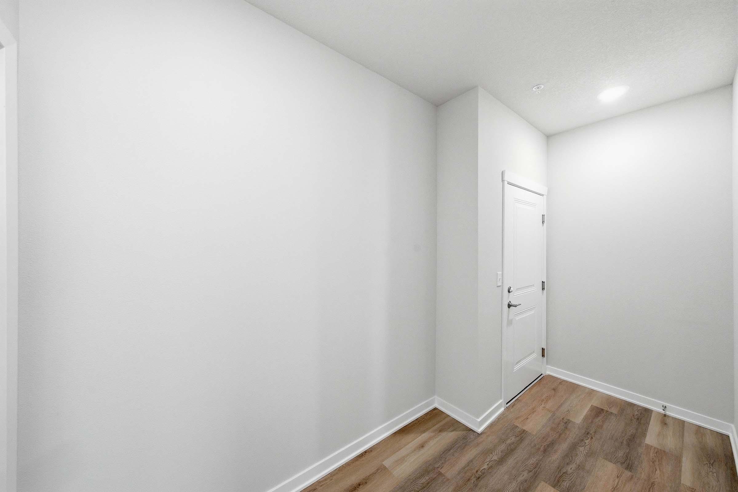 A well-lit, empty hallway with light-colored walls and a wooden floor. The space features a closed door on the right side, and the opposite wall is clear, creating a simple and uncluttered appearance.