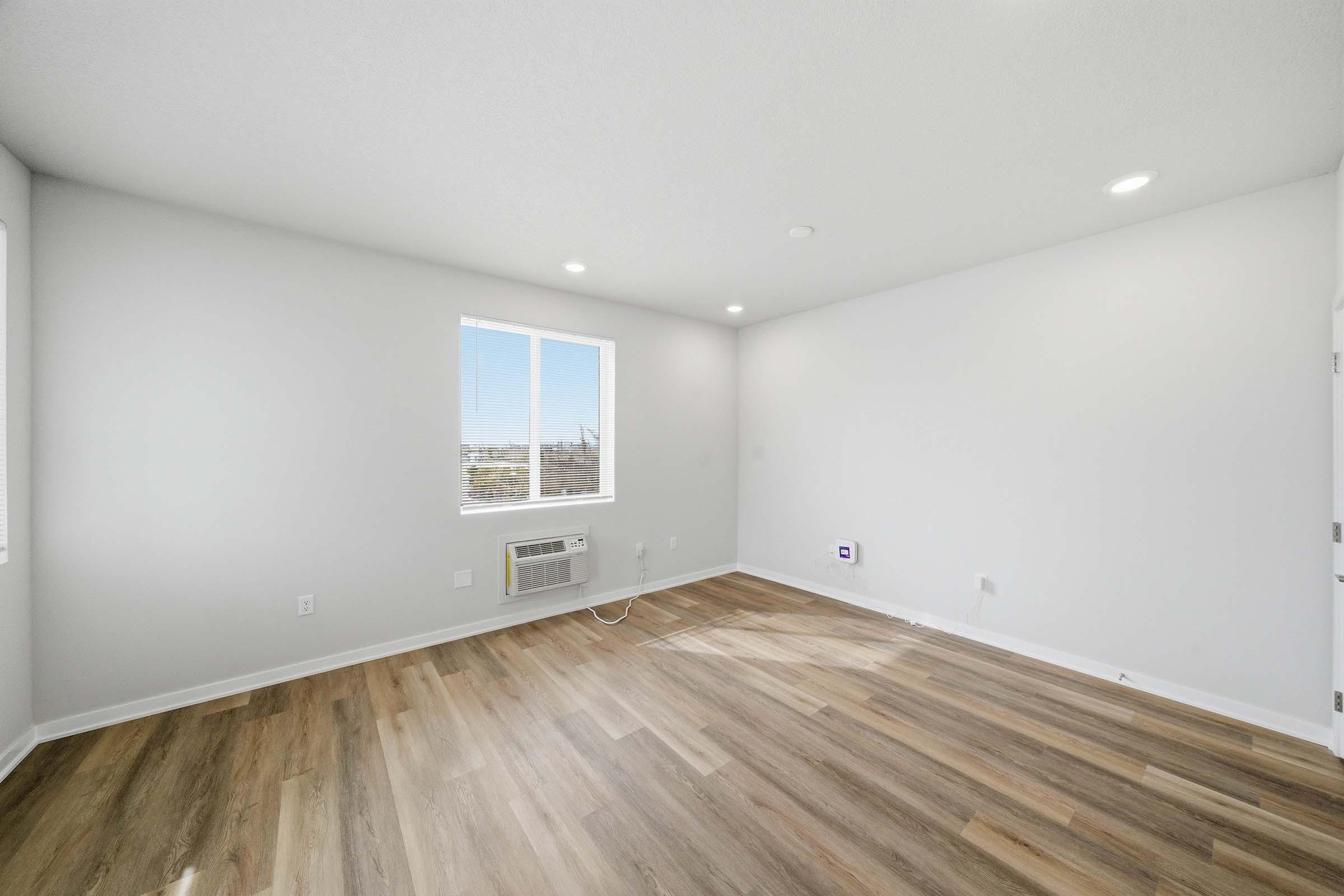 A bright, unfurnished room featuring light-colored walls and a hardwood floor. A window allows natural light to enter, and there is an air conditioning unit mounted on the wall. The space is minimalistic and ready for furnishing.