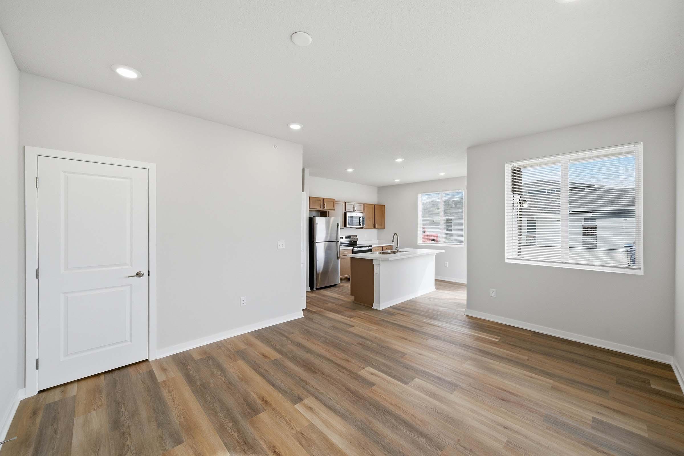 Bright and modern open-concept living space featuring light wood flooring, neutral walls, and a large window providing natural light. The kitchen is equipped with stainless steel appliances and has a countertop with seating. A door leads to a separate room, possibly a bedroom or bathroom.