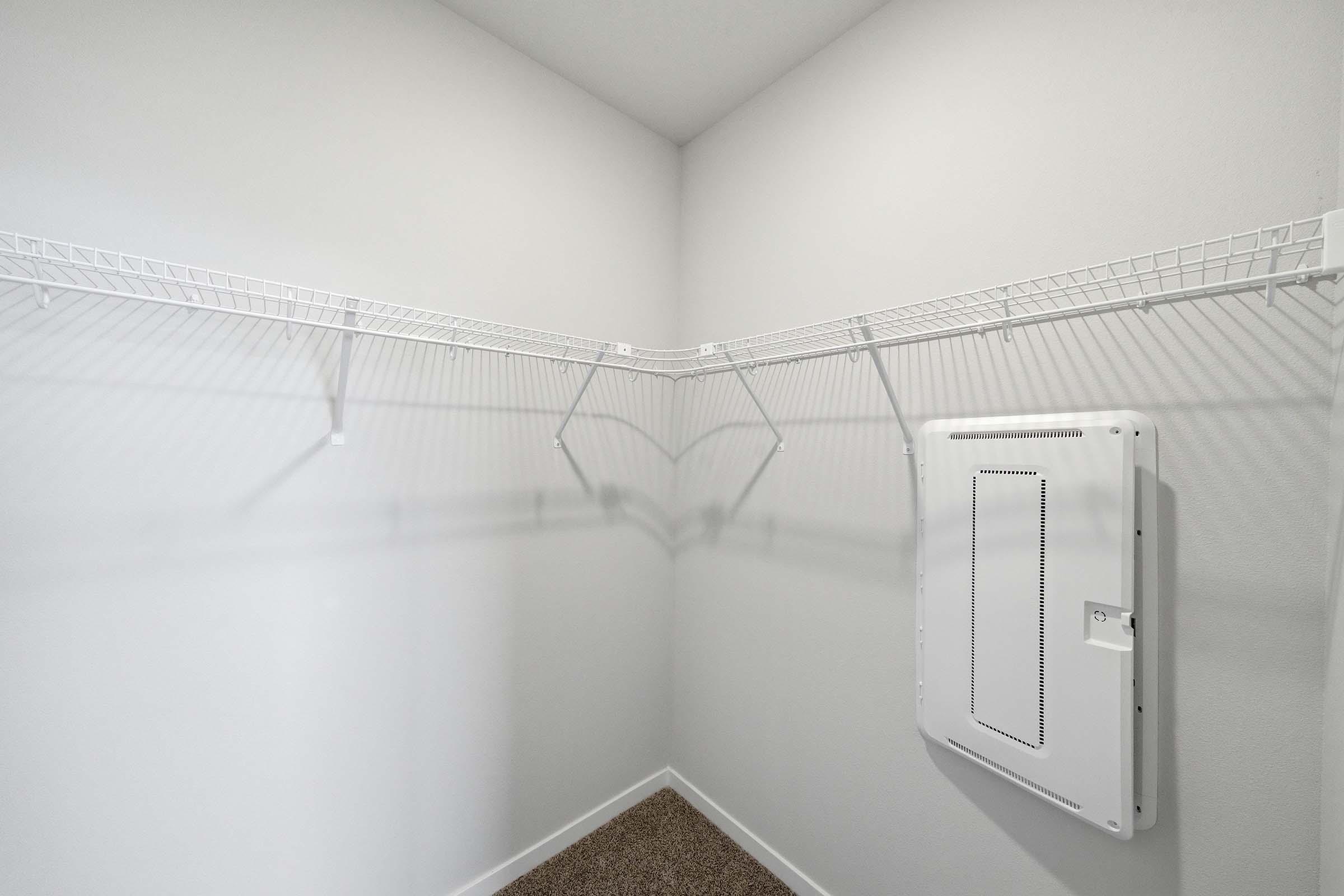 An empty corner closet with white wire shelving installed along the walls. The walls are painted light gray, and there is a small white vent or panel on one wall. The floor is covered with a tan carpet.