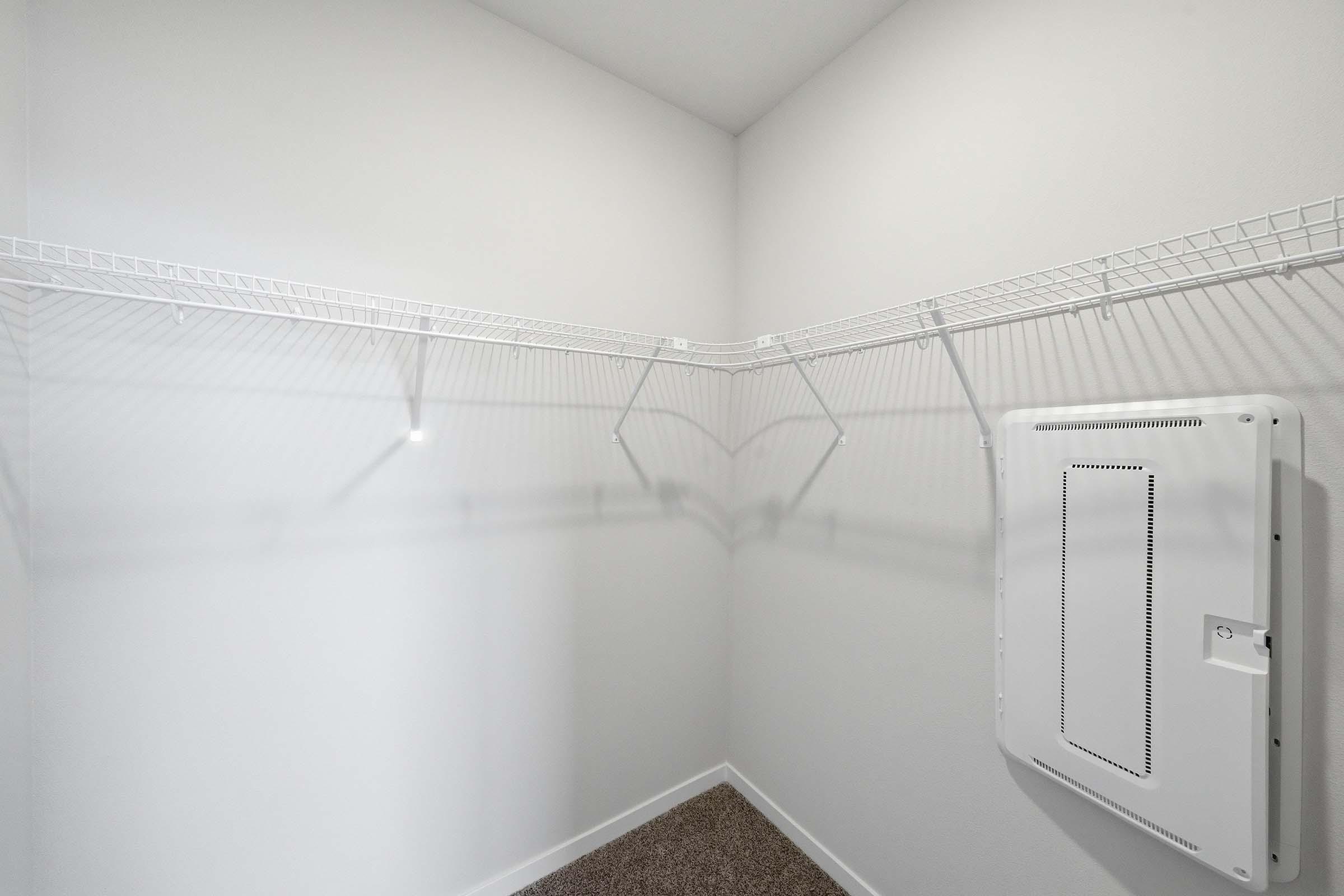 Empty walk-in closet with white walls, featuring two wire shelves along the corner and a heating unit mounted on the wall. The carpeted floor is visible, enhancing the spacious feel of the room.