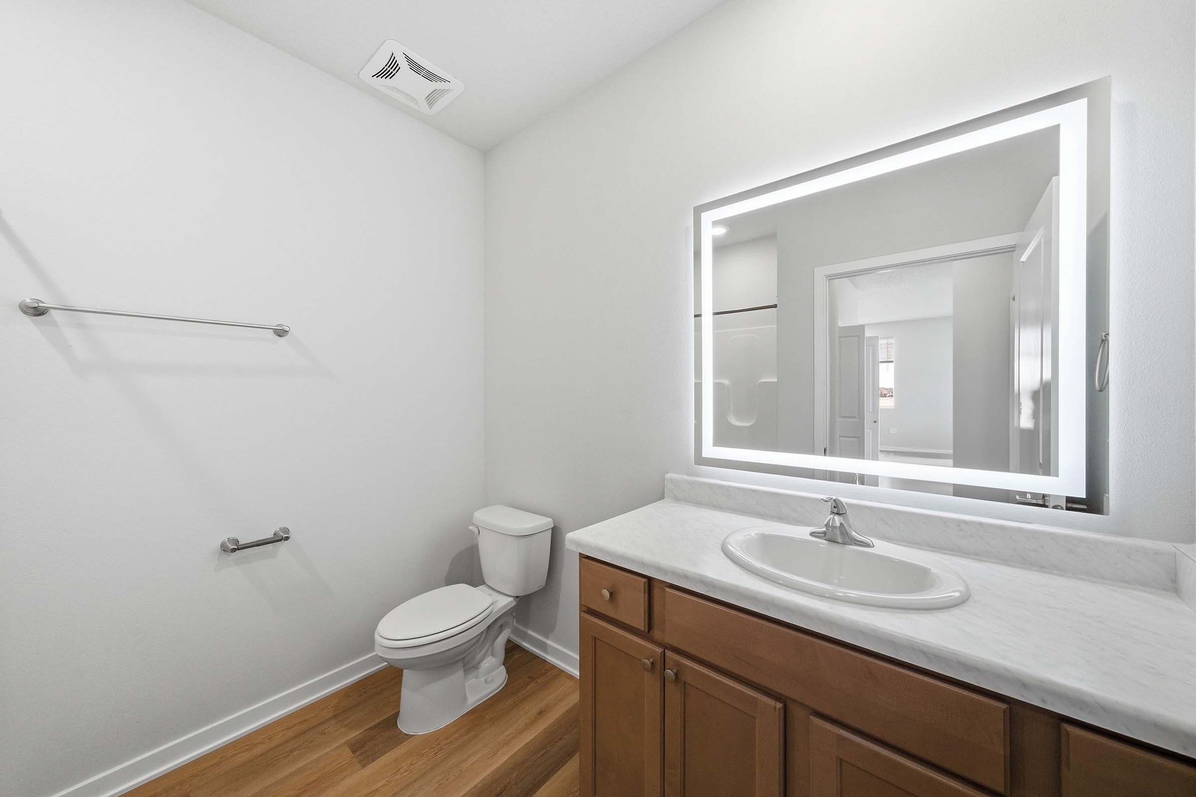 A clean and modern bathroom featuring a white wall, laminate flooring, a toilet, and a vanity with a sink. A large illuminated mirror enhances the space, and a towel bar is mounted on the wall. The design is minimalist and functional, emphasizing bright lighting and simplicity.