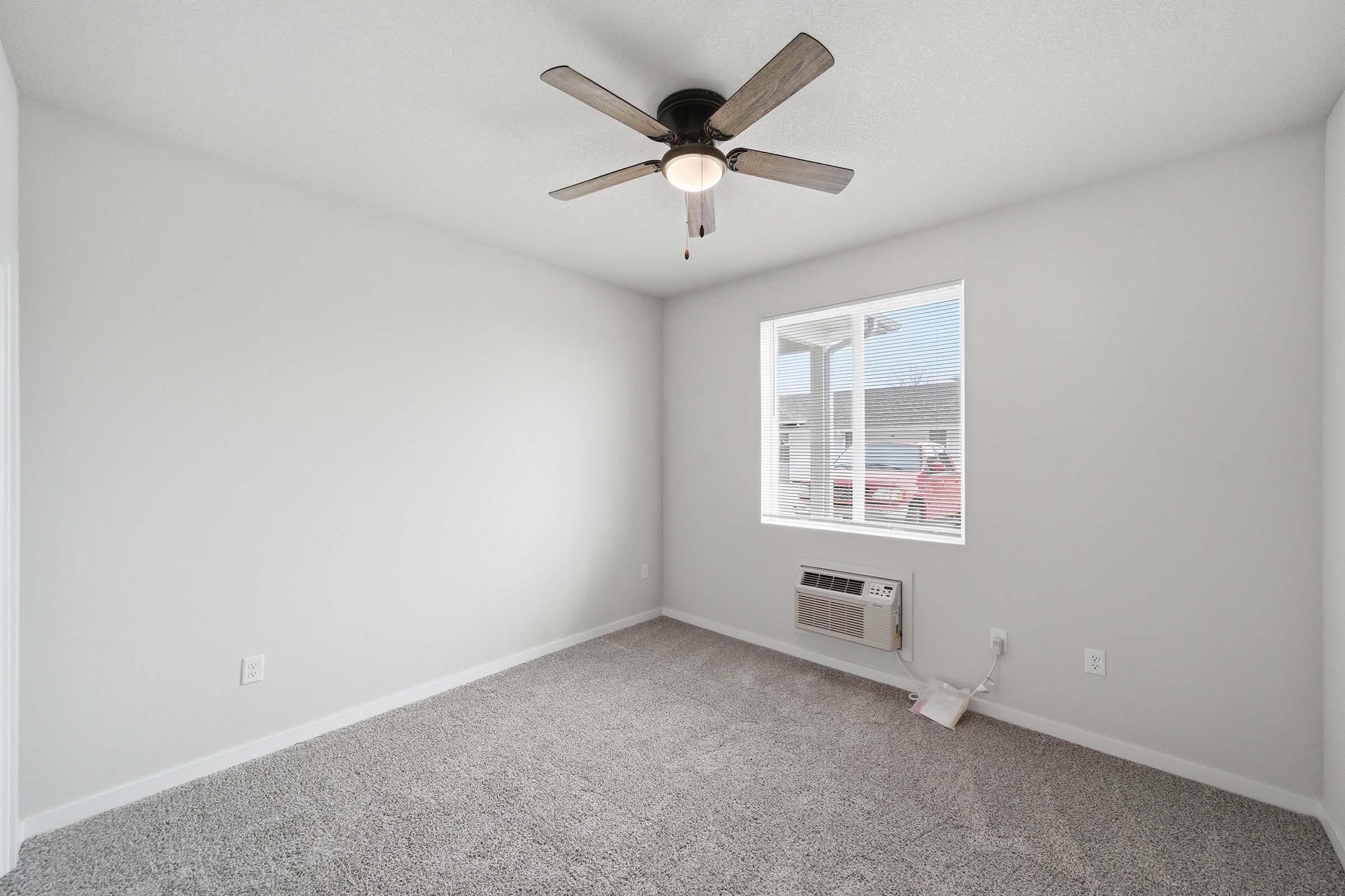 A vacant room featuring light gray walls, plush gray carpet, and a ceiling fan. There's a window allowing natural light and an air conditioning unit on the wall. The room has a simple and minimalistic design, making it suitable for various purposes.