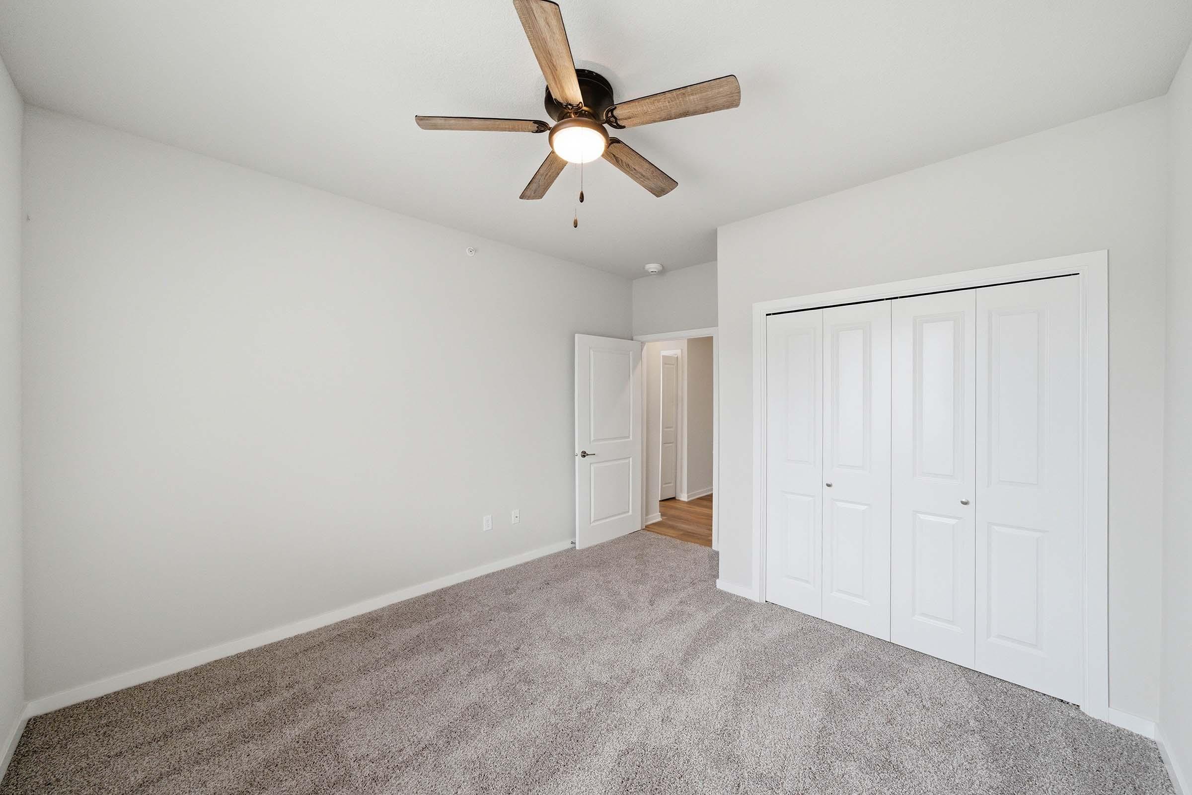 A spacious, empty bedroom featuring light gray walls and plush carpet. It has a ceiling fan with light, a closet with sliding doors, and an entrance to another room visible in the background. Natural light brightens the space, creating a calm and inviting atmosphere.
