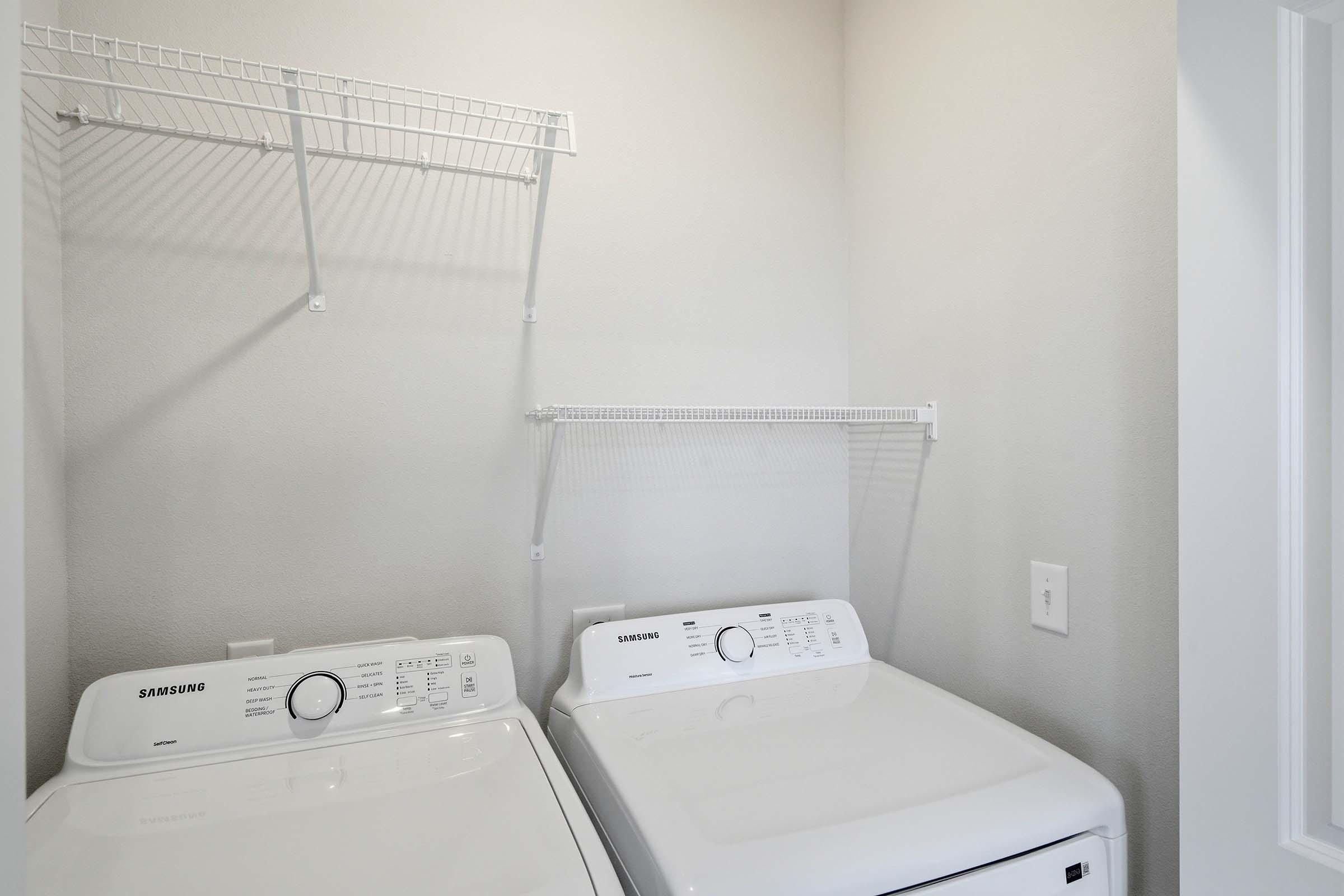 A clean laundry room featuring two Samsung washing machines side by side. Above them, a wire shelving unit provides storage space. The walls are painted a light color, creating a bright and spacious feel in the room.