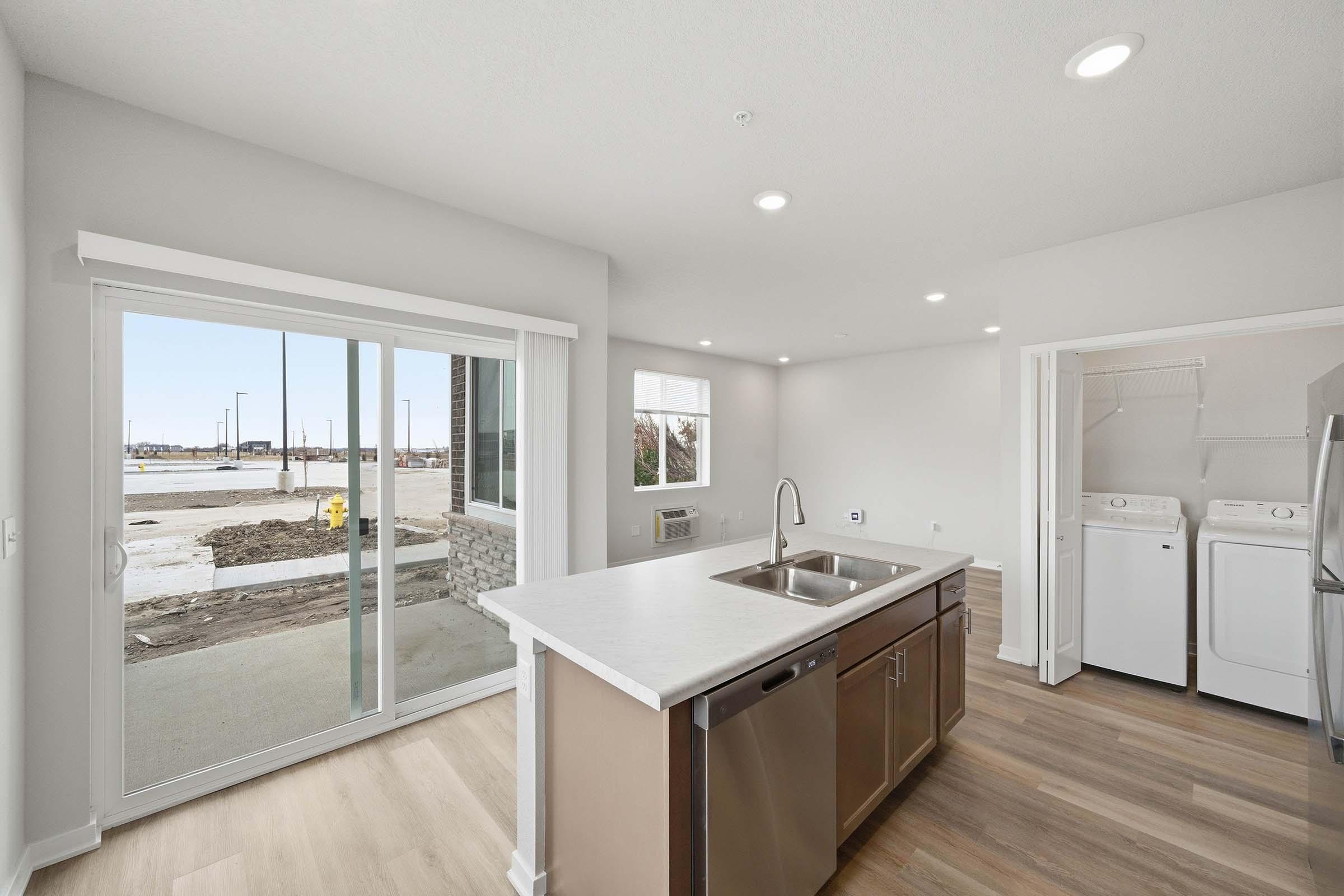 Modern kitchen and laundry area with light wood flooring, white walls, and a large window. The kitchen features a central island with a sink and stainless steel appliances. There is a sliding glass door leading to an outdoor area under construction. Natural light brightens the space.