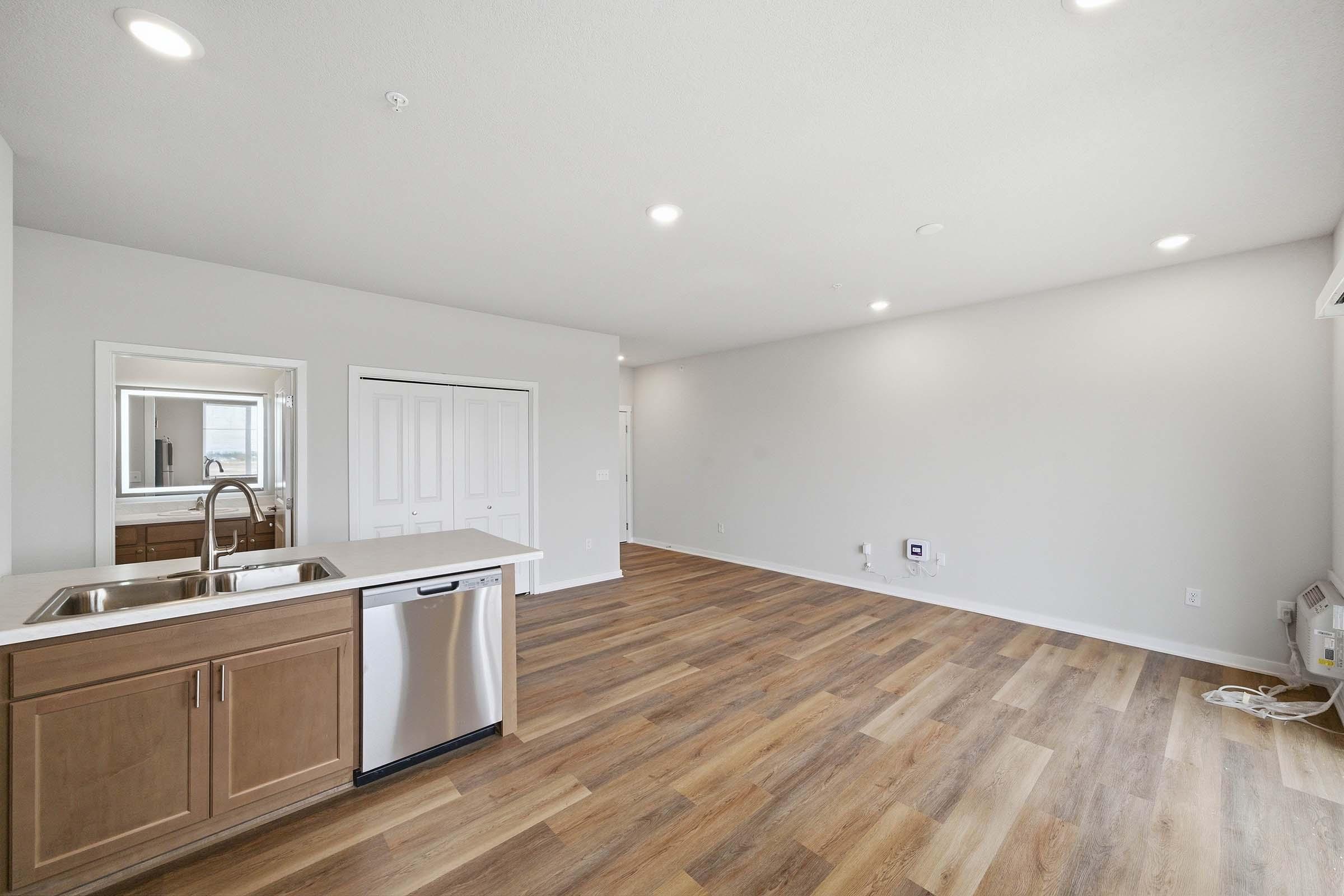 A bright, spacious living area featuring light-colored walls and oak laminate flooring. On the left, a modern kitchen with wooden cabinets and a stainless steel dishwasher is visible. The open layout promotes a seamless flow into the rest of the room, which has natural light and ample space.