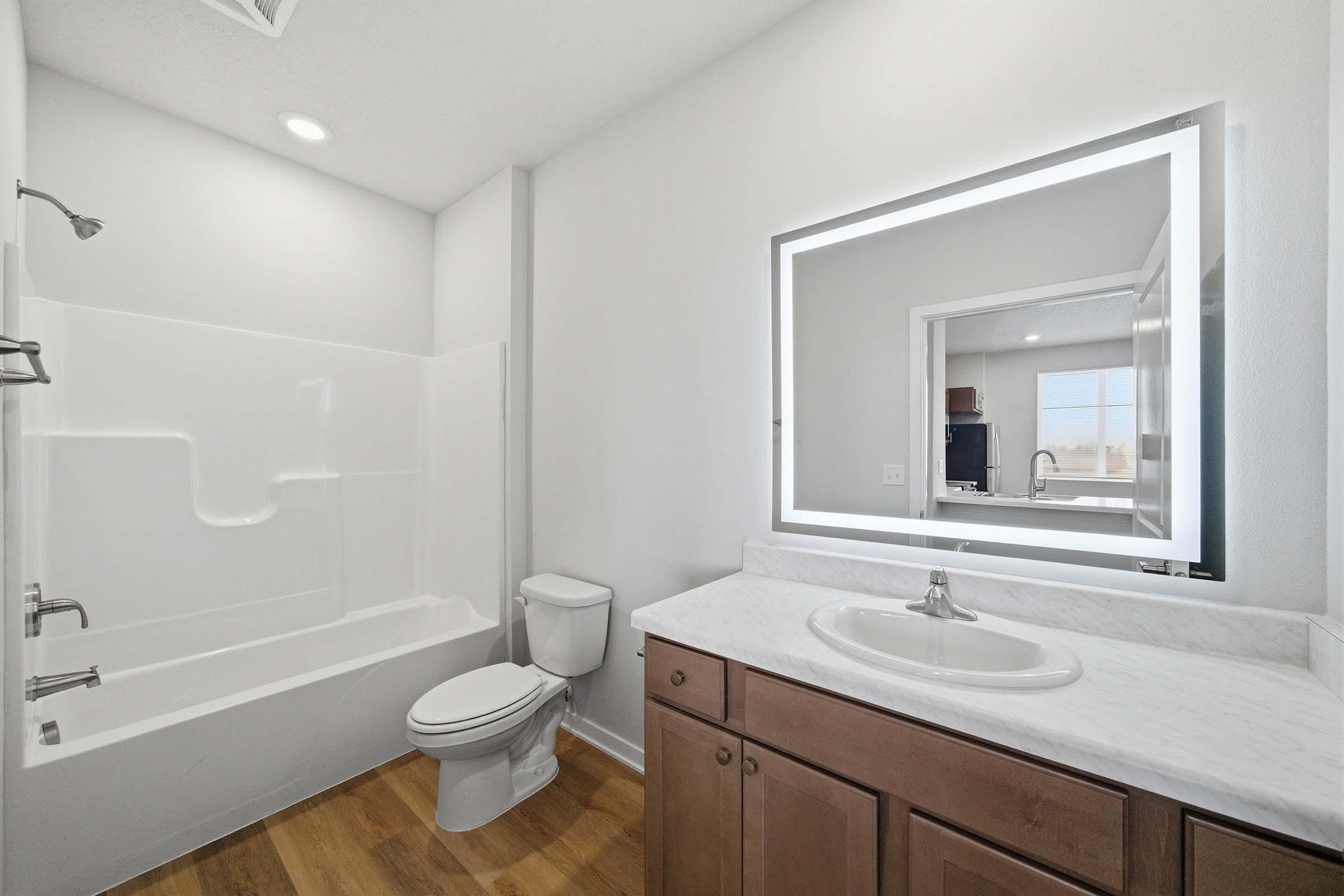 A modern bathroom featuring a white bathtub and shower combination, a toilet, and a large mirror with integrated lighting above a white countertop sink. The walls are painted white, and there is a light wooden floor. Natural light is visible through a window in the background.