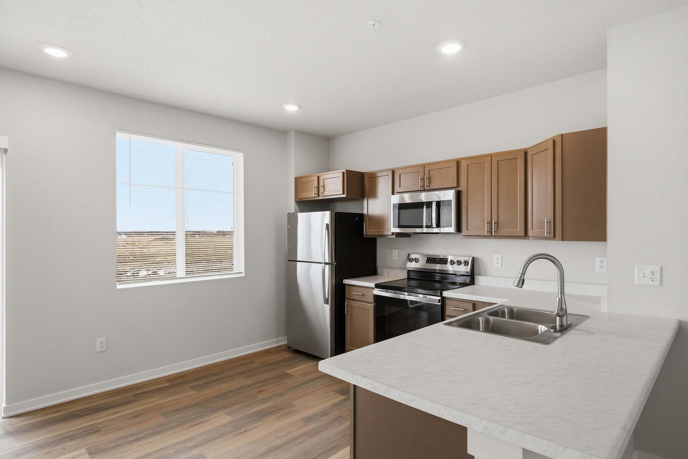 Modern kitchen with light gray walls and sleek wooden cabinets. Features stainless steel appliances, including a refrigerator, oven, and microwave. A white marble countertop with a sink is visible. Large window allows natural light to brighten the space, highlighting the wood flooring.
