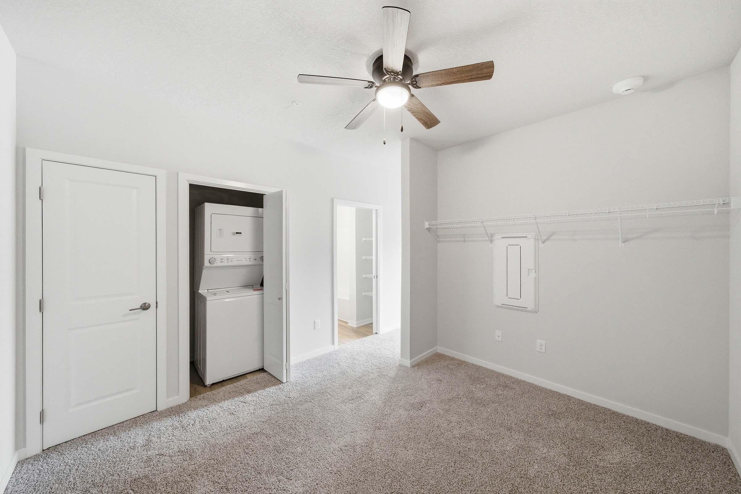 A spacious, empty bedroom with light-colored walls and carpet. It features a ceiling fan and a small laundry area with washer and dryer visible in the doorway. A sliding door leads to a storage area, while a wire shelf is mounted on one wall, providing extra space for storage or organization.