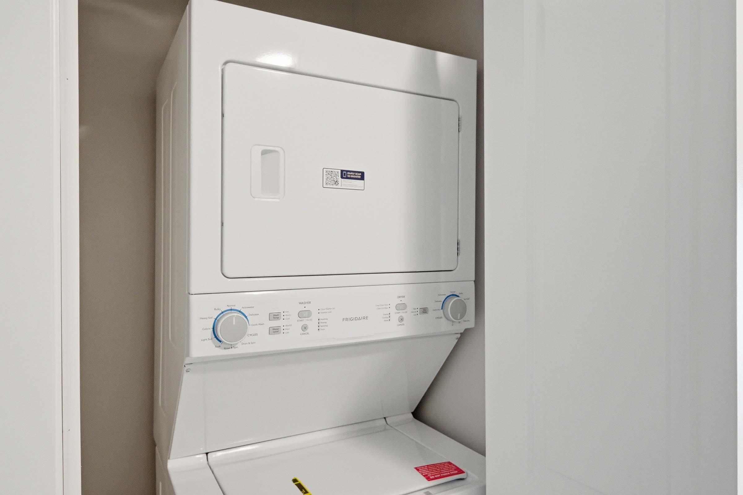 A stacked washer and dryer unit in a small laundry closet, featuring a white finish and control dials at the front. The dryer is positioned above the washer, both appliances have a modern design. There is a warning label on the washer's door. The interior is well-lit and minimalistic.