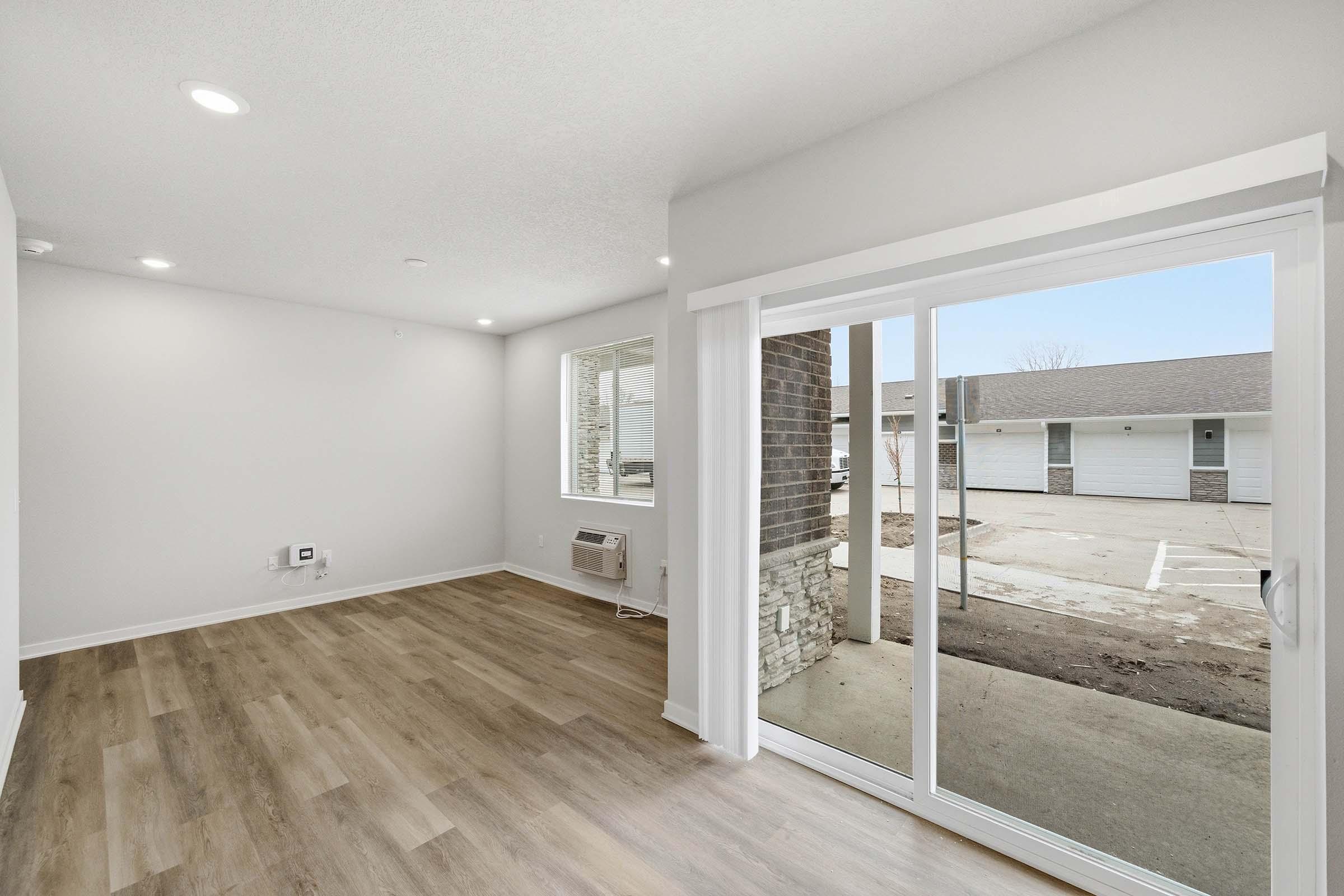 A modern, empty room featuring light-colored walls and wood-style flooring. Large windows allow natural light to fill the space, and there's a sliding glass door leading outside. The view includes a parking area with several white buildings in the background.