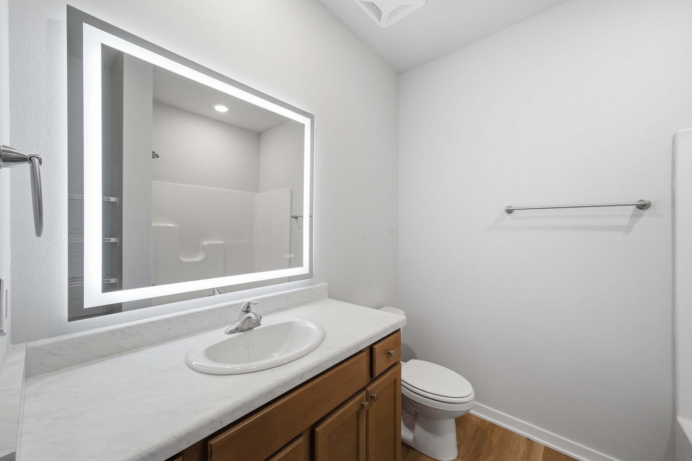 A modern bathroom featuring a white countertop with a circular sink, a large illuminated mirror, light-colored walls, and a wooden floor. The space includes a toilet and a towel rack, with a glass shower stall partially visible in the background.