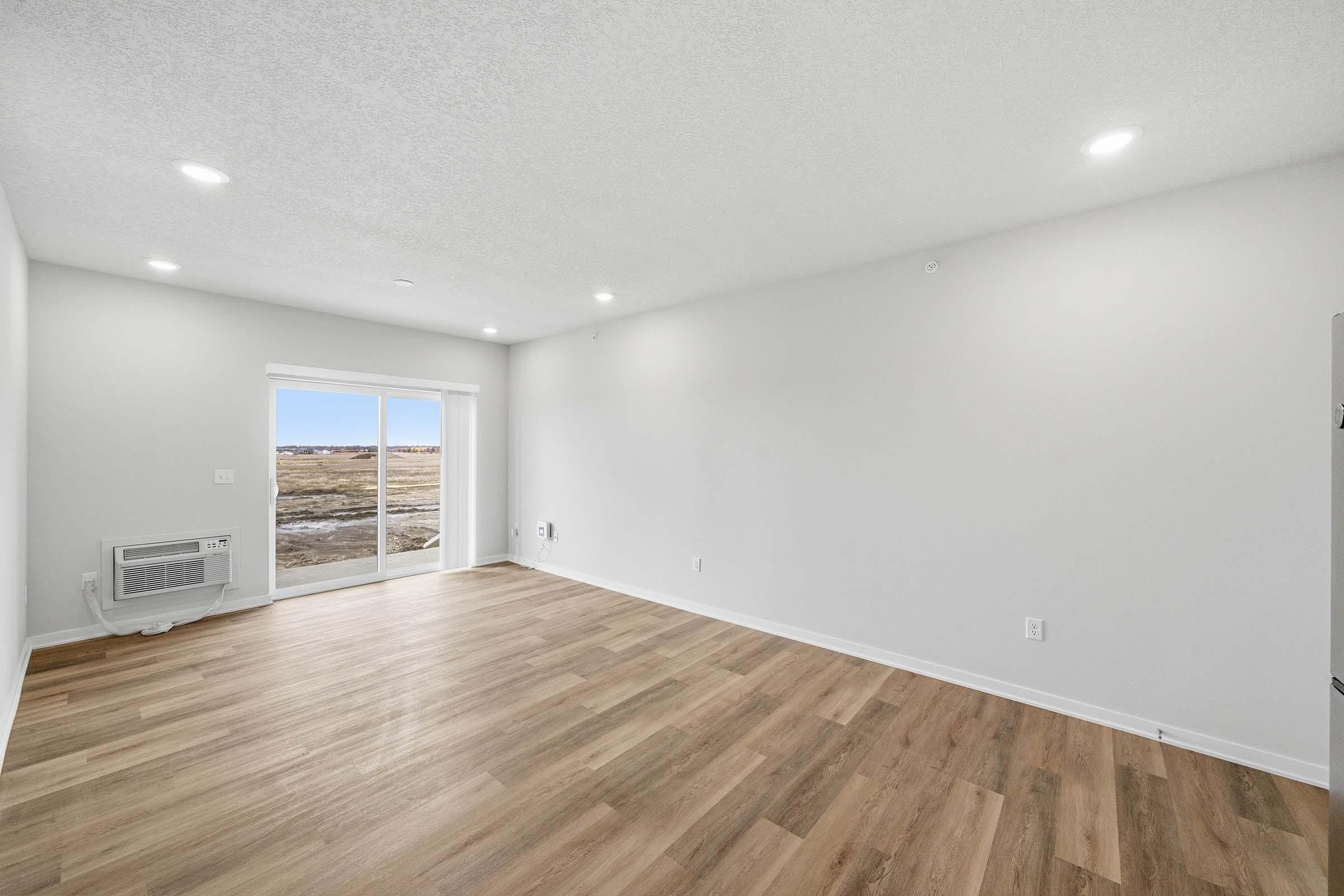 A bright and spacious empty living room with light-colored walls and wooden flooring. Large sliding glass doors open to an outside view, allowing natural light to enter. There’s an air conditioning unit on the left wall, and the space is ready for furniture arrangement.
