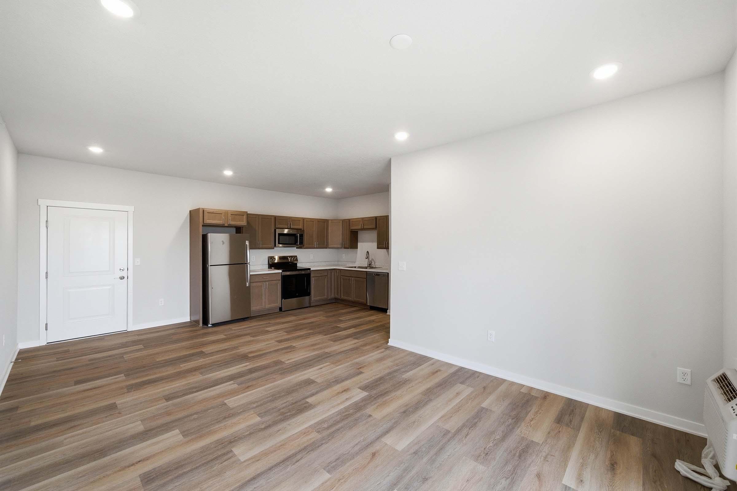 A modern, open-concept kitchen and living area featuring light-colored walls, wooden flooring, and ample natural light. The kitchen includes stainless steel appliances, wooden cabinets, and a spacious layout, while the living area offers plenty of room for furniture arrangements.