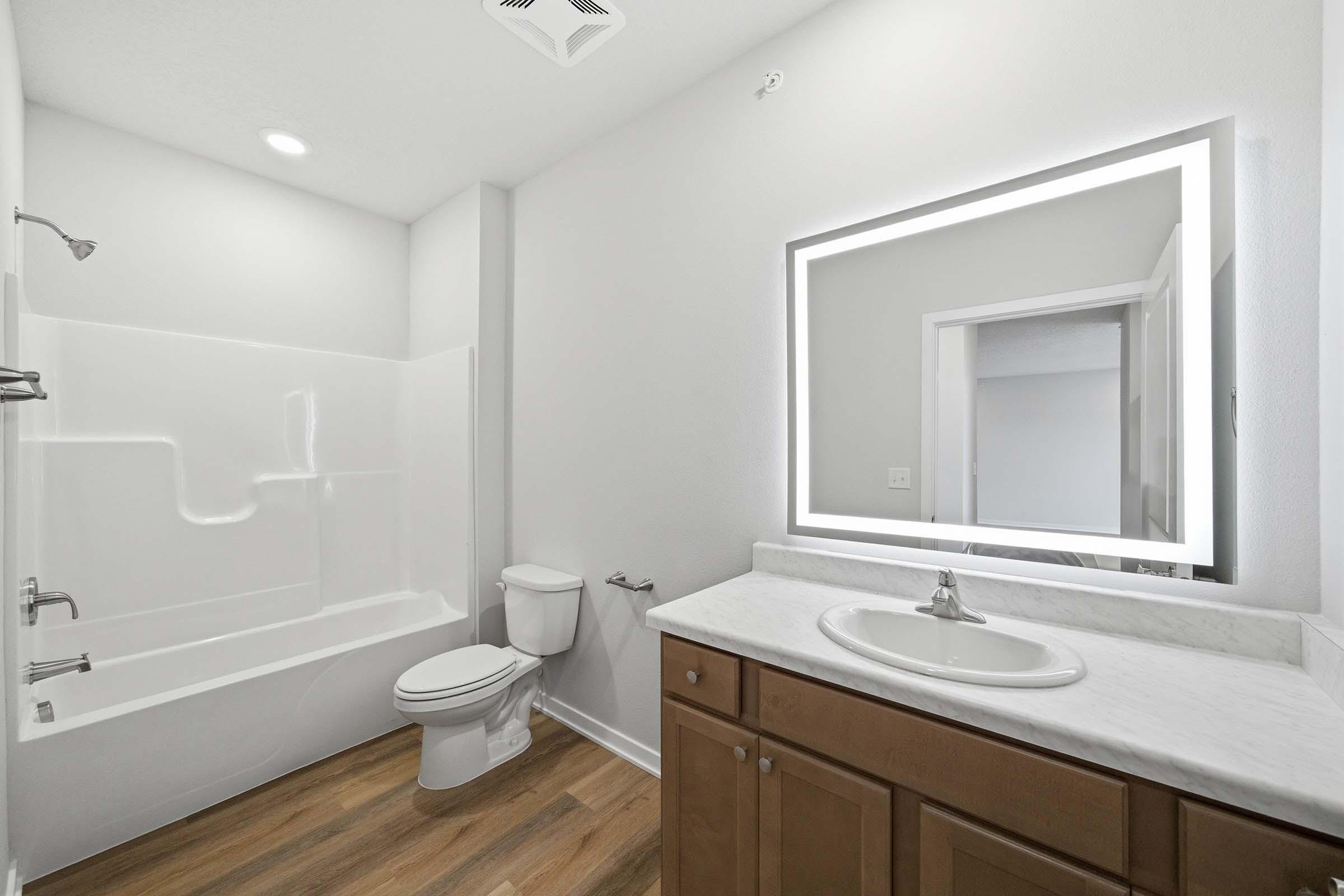 A modern bathroom featuring a bathtub with a shower, toilet, and a vanity with a marble countertop. The space has light-colored walls and wood laminate flooring. A large, illuminated mirror hangs above the sink, adding a contemporary touch to the overall design.