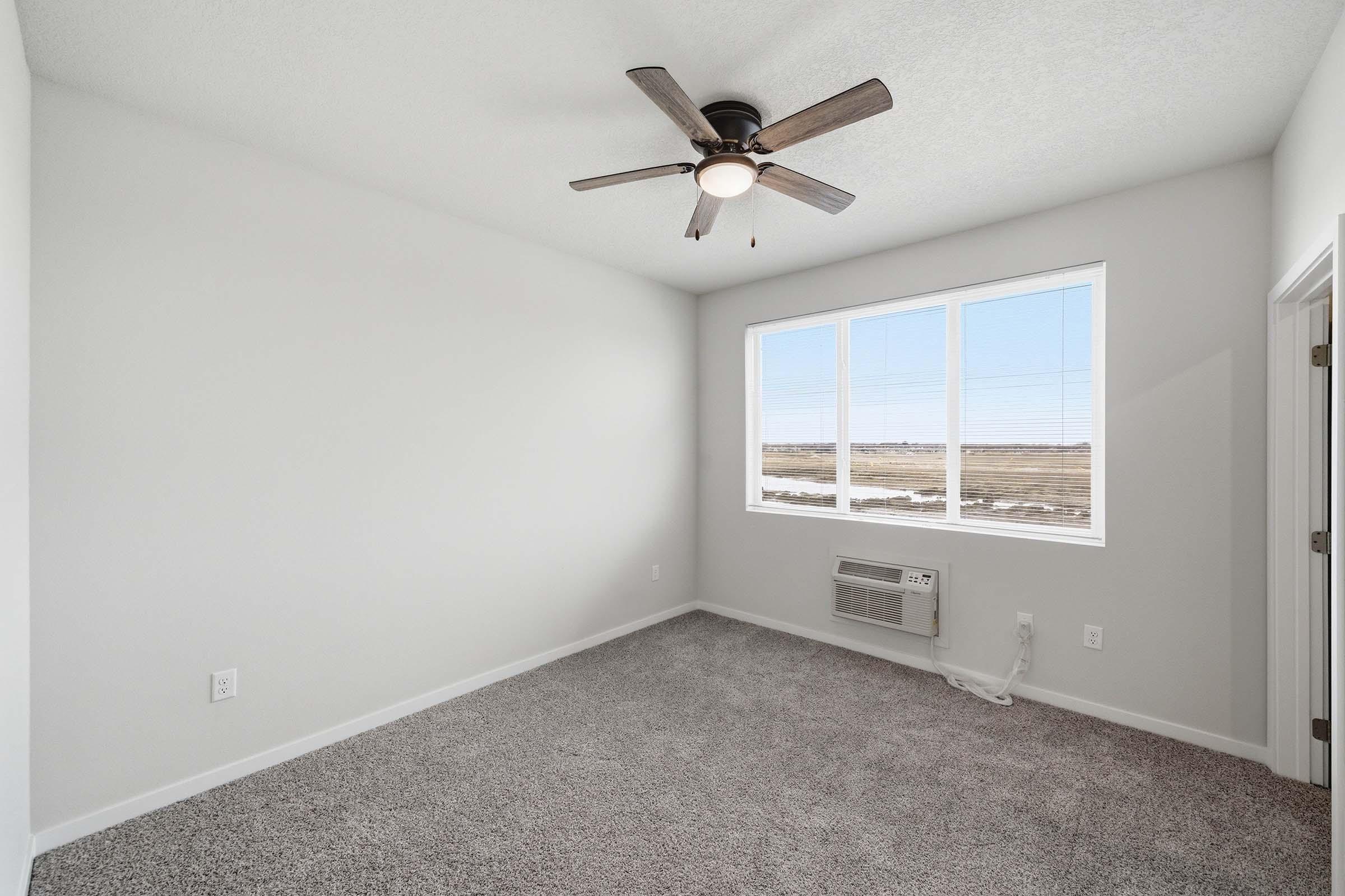 A bright, empty room with gray carpet and white walls. It features a ceiling fan and a large window that offers a view of the outdoor landscape. An air conditioning unit is visible on the wall under the window.