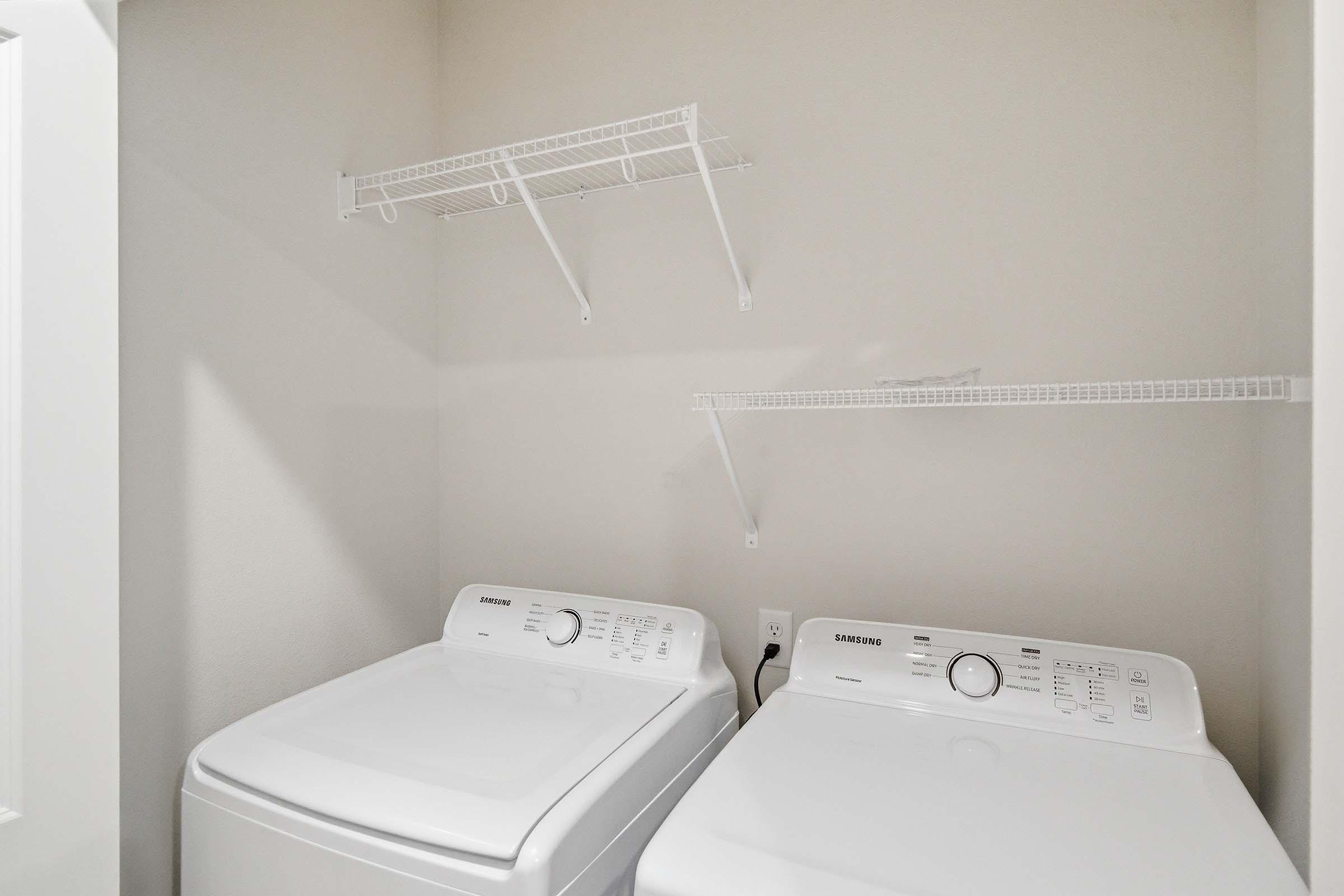 A laundry room featuring two modern washing machines side by side, with two white wire shelves mounted on the wall above. The walls are painted in a light color, enhancing the spacious and clean look of the area.