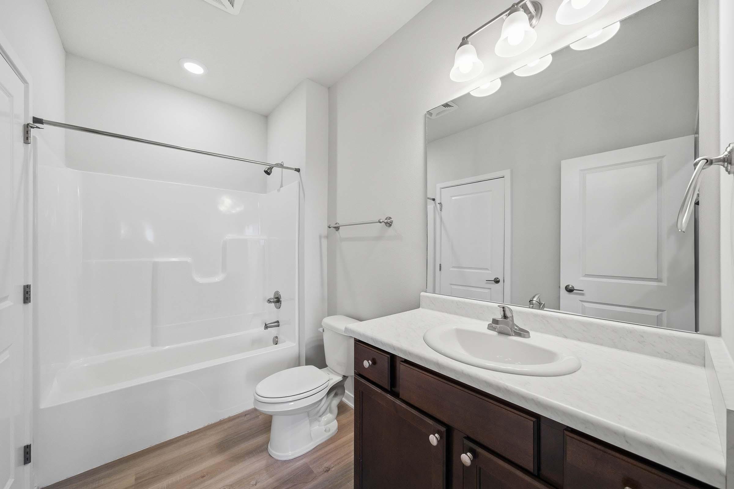 A modern bathroom featuring a clean design with a white bathtub and shower combination, a simple countertop with a sink, and dark wooden cabinetry. There is a toilet next to the sink, and the walls are painted light grey. Bright overhead lighting enhances the spaciousness of the room.