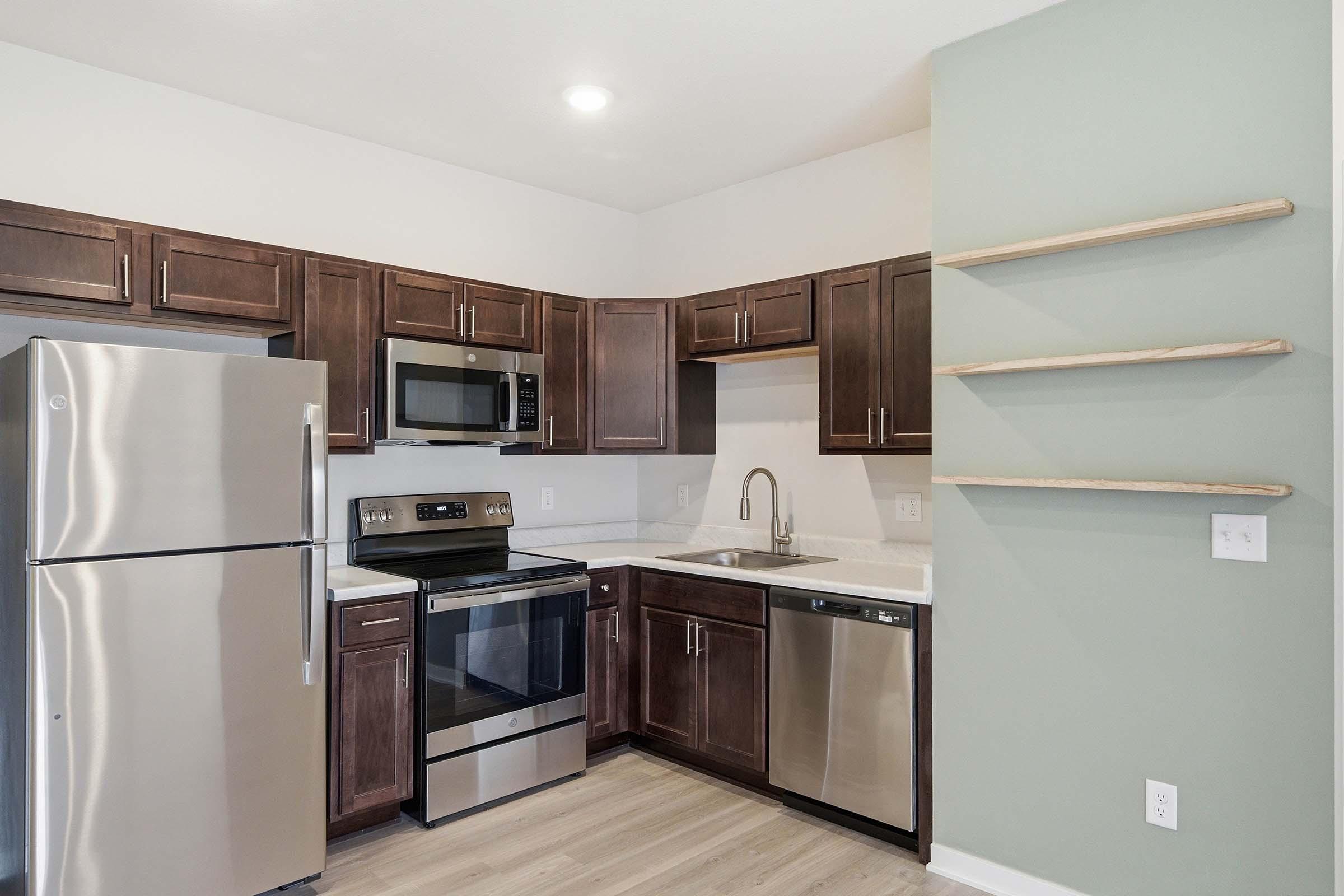 Modern kitchen featuring stainless steel appliances, including a refrigerator, microwave, and oven. The cabinetry is dark wood, complemented by a light countertop. A wall-mounted shelf displays a minimalist design on one side. The flooring is light-colored, adding to the contemporary aesthetic.