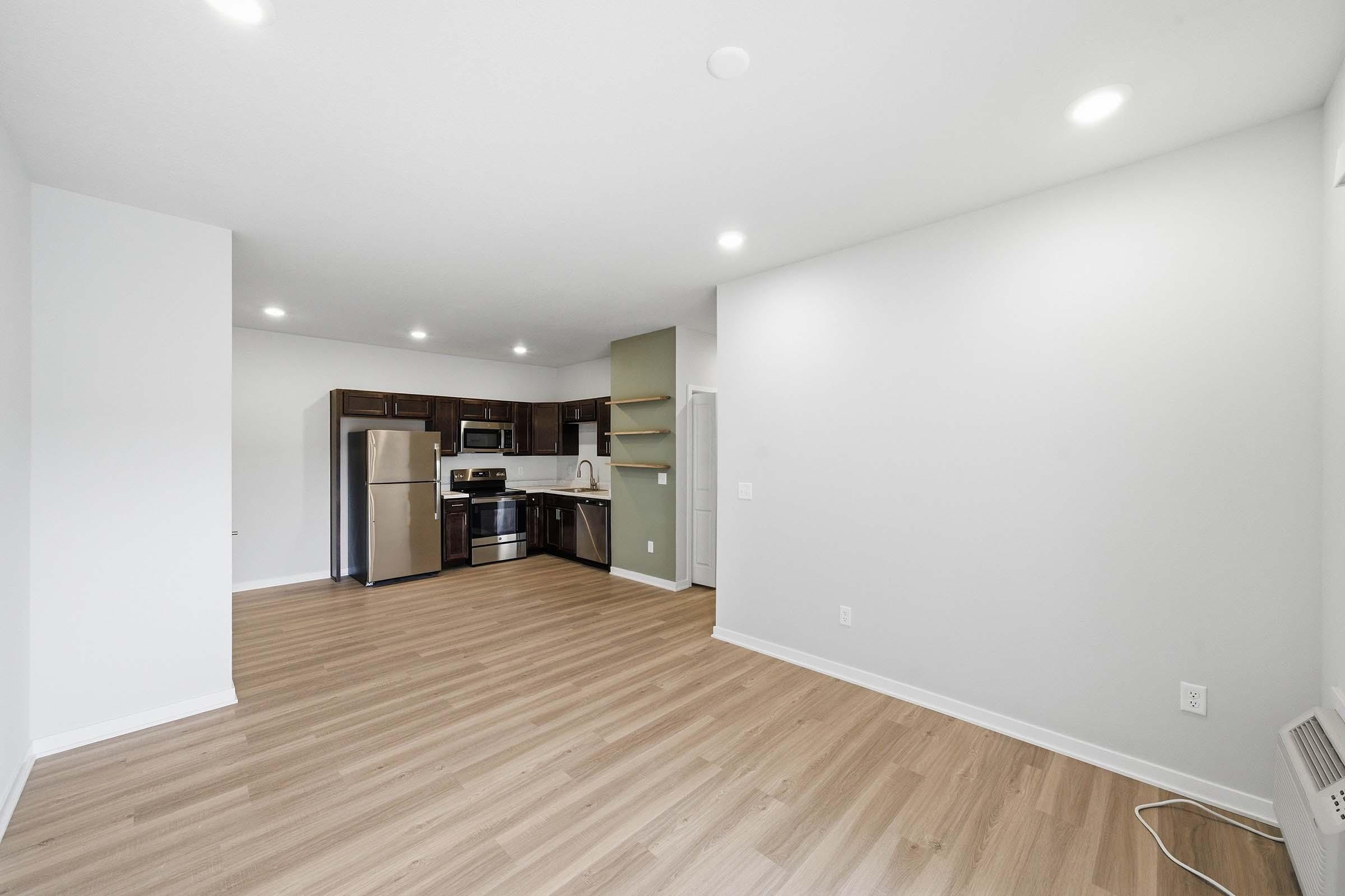 A spacious, modern living area with light-colored wood flooring. The kitchen features dark cabinets, stainless steel appliances, and an open layout. Bright overhead lights illuminate the space, which is painted in neutral tones, creating a welcoming atmosphere. A door leads to another room, enhancing the sense of openness.