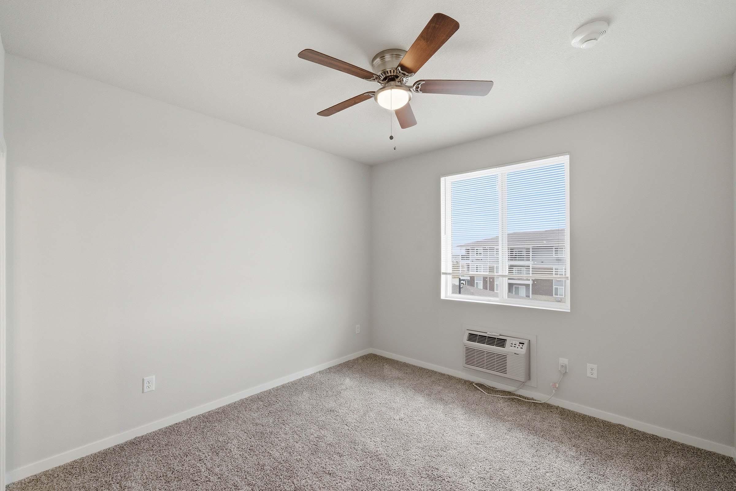 A well-lit, empty bedroom featuring light-colored walls and plush carpet. The room has a ceiling fan with wooden blades, a window with blinds allowing natural light, and an air conditioning unit on the floor. The space appears clean and ready for new furnishings.