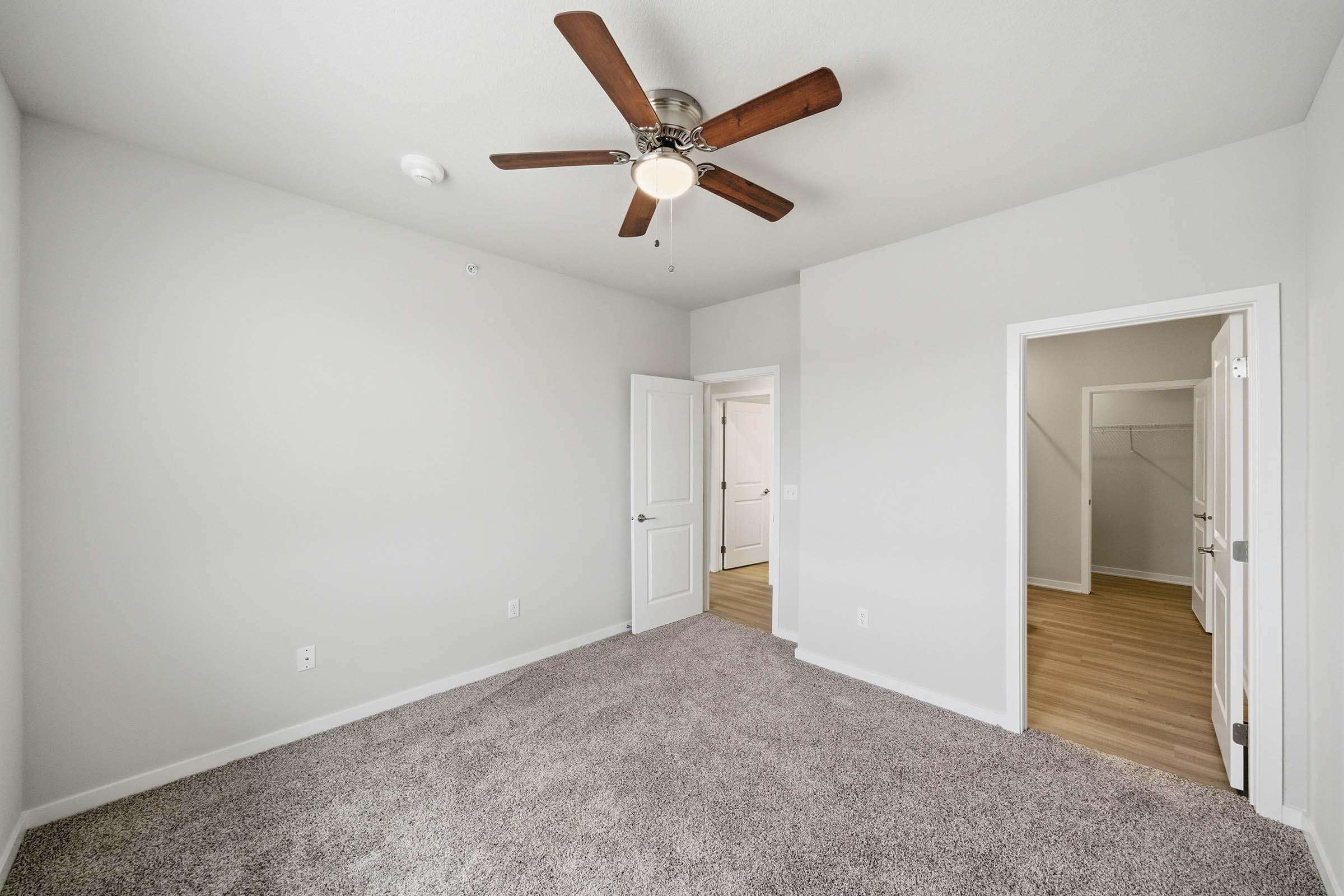 A spacious, neutral-colored bedroom featuring light gray walls and a plush carpet. A ceiling fan with wooden blades hangs from the ceiling. To the right, a doorway leads to a closet or another room with hardwood flooring. The overall ambiance is clean and modern, suitable for various decor styles.