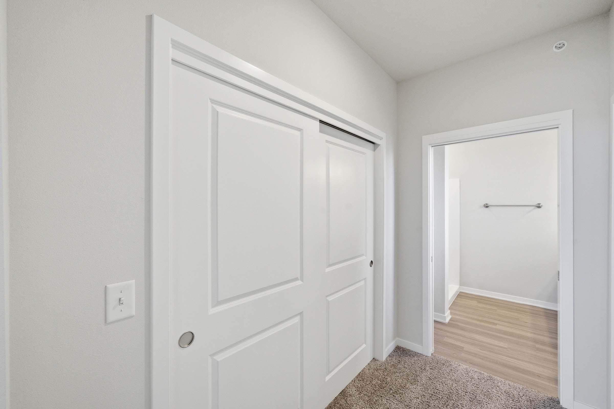 A well-lit hallway featuring a sliding white door on the left and an open doorway leading to another room on the right. The walls are light-colored, and the floor has a soft carpet transitioning to a smooth wood finish in the adjacent room, creating a clean and modern aesthetic.