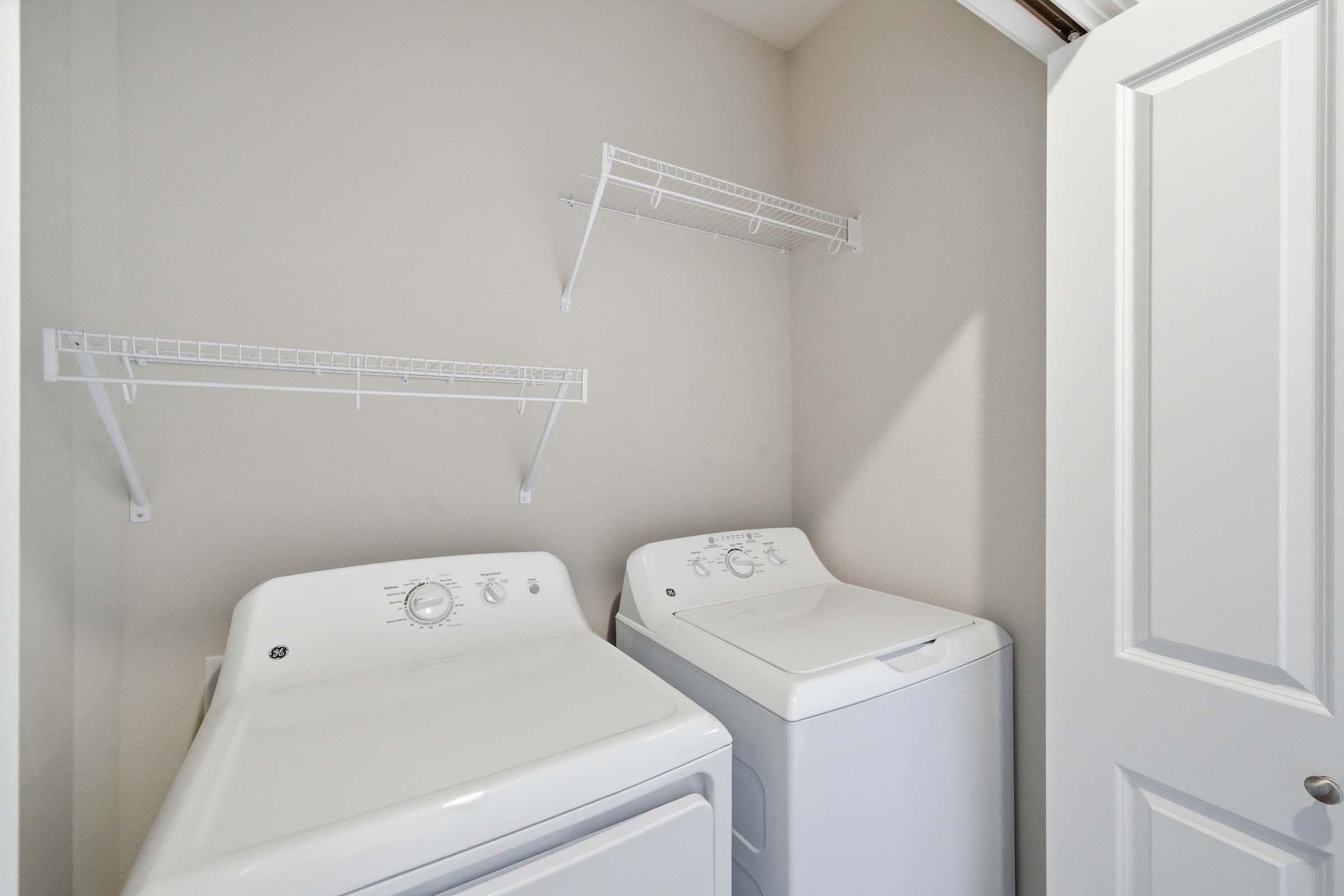 A small laundry room featuring two white washing machines side by side, with wall-mounted wire shelves above them. The room has light-colored walls and a door partially closed, creating a tidy and organized appearance.
