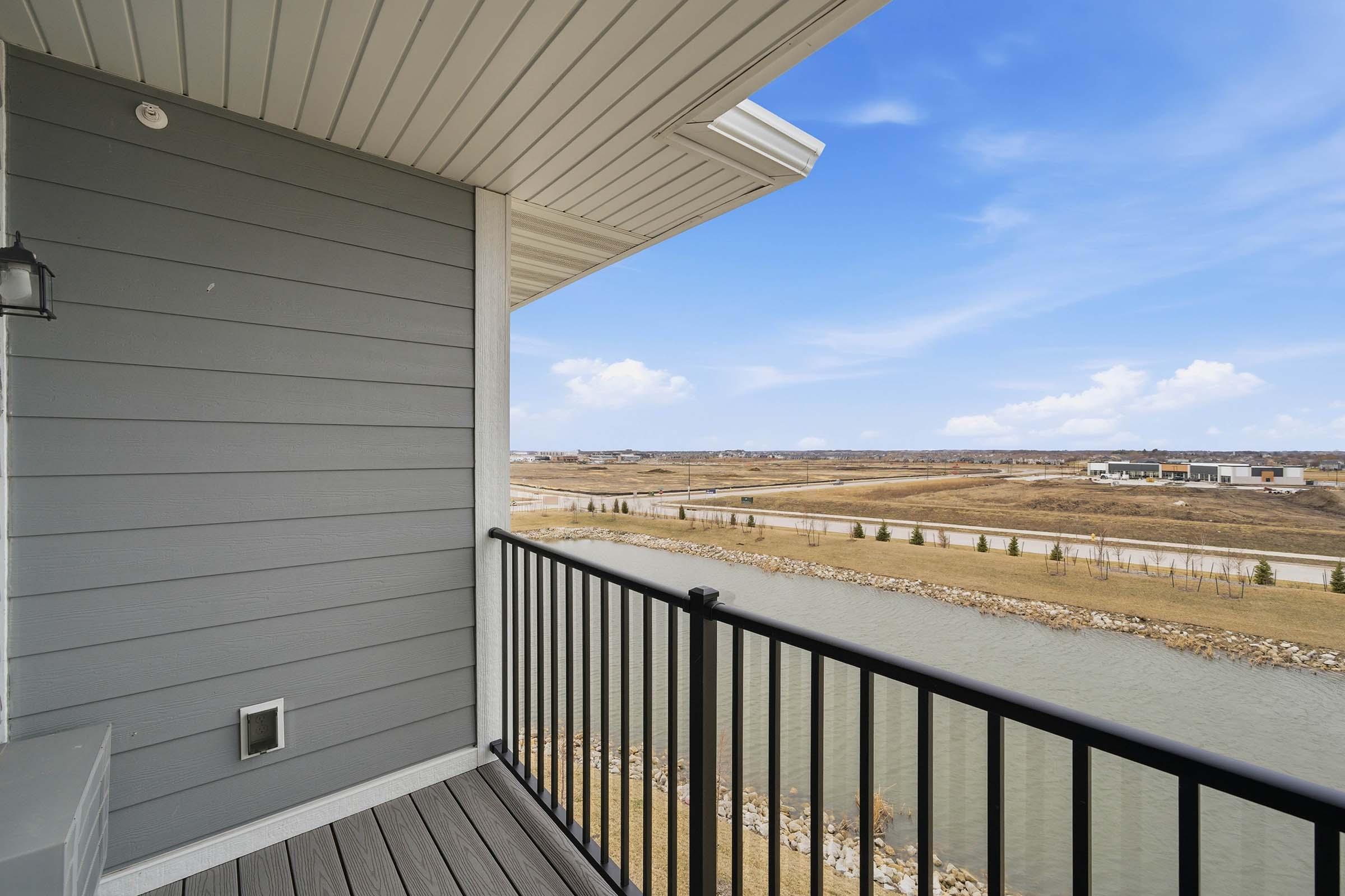 View from a balcony featuring a railing, overlooking a calm body of water and a landscape with rolling hills. The sky is clear with a few clouds, and there are buildings in the distance along the shore. The setting appears serene and is situated in a developed area.