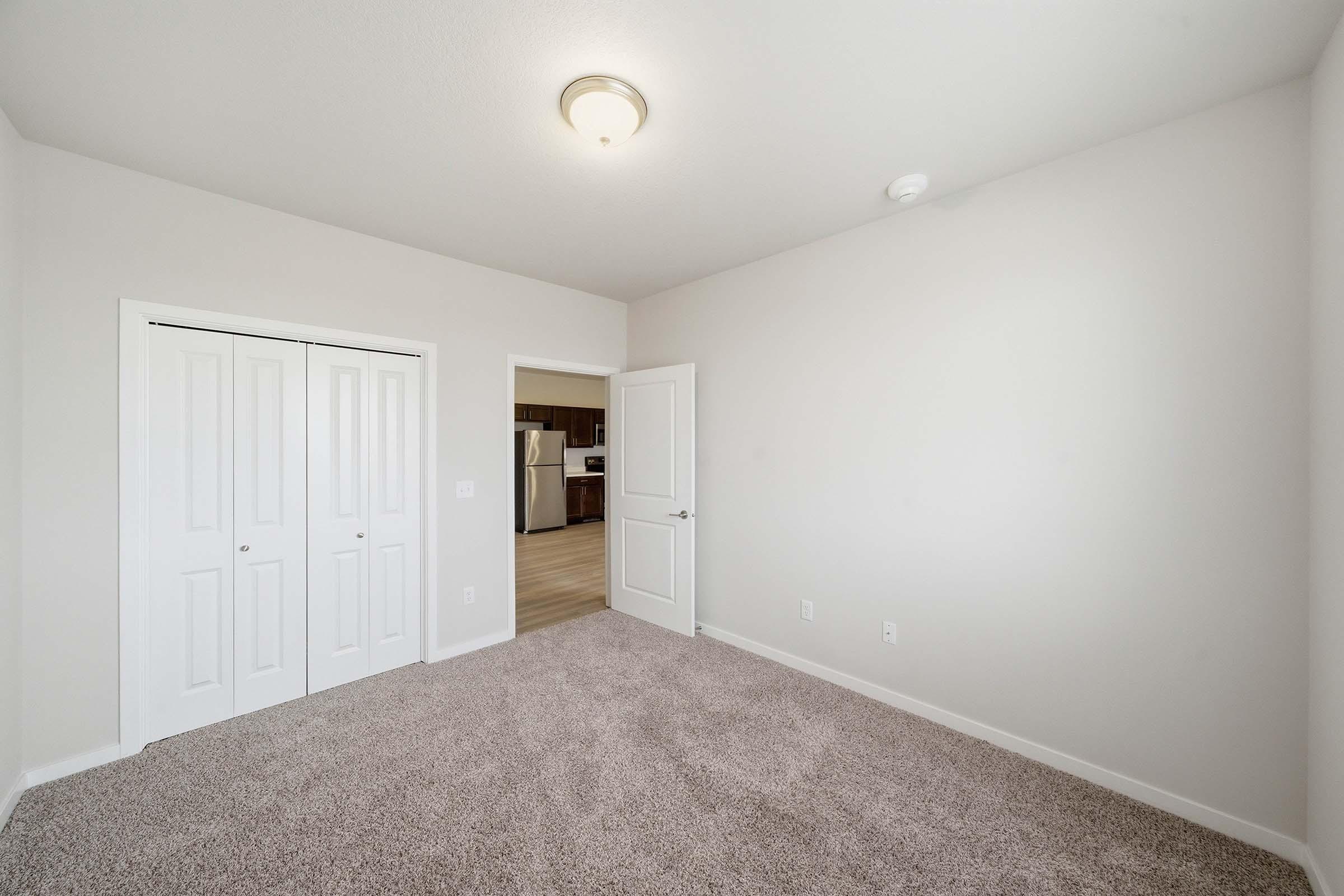 A vacant room with beige walls and light-colored carpet, featuring a ceiling light fixture. There are double white closet doors on the left and a door leading to another room on the right. The space is well-lit and appears spacious, with minimal furnishings.