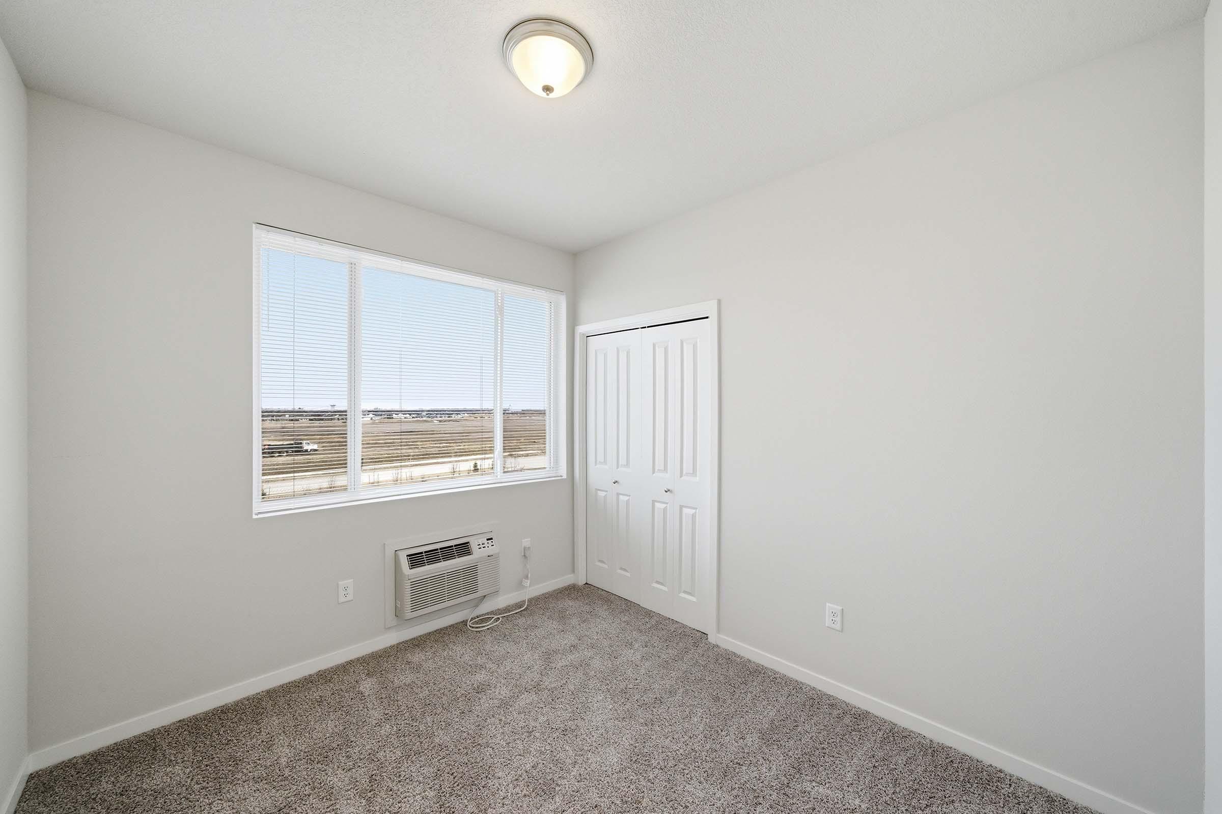 A bright, empty room with light-colored walls and carpet. It features a large window with blinds, an air conditioning unit, and a closet with double doors. The view outside shows a clear sky and open landscape. The overall atmosphere is clean and spacious, suitable for various uses.
