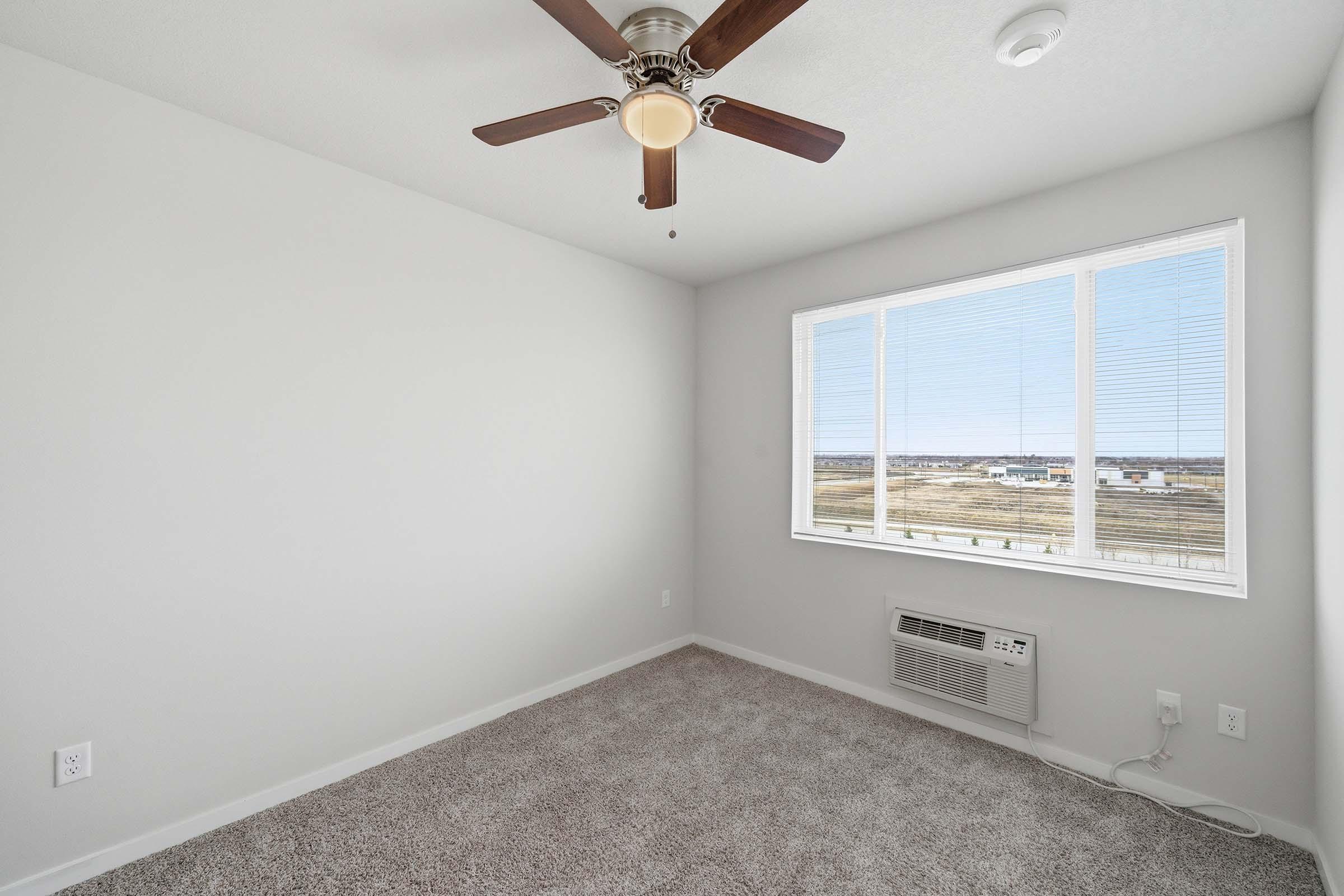A bright, empty room with light gray walls and a ceiling fan. It features a large window with blinds, showing a view of open space outside. The floor is covered with soft, beige carpet, and there's a wall-mounted air conditioning unit. The overall atmosphere is spacious and well-lit.