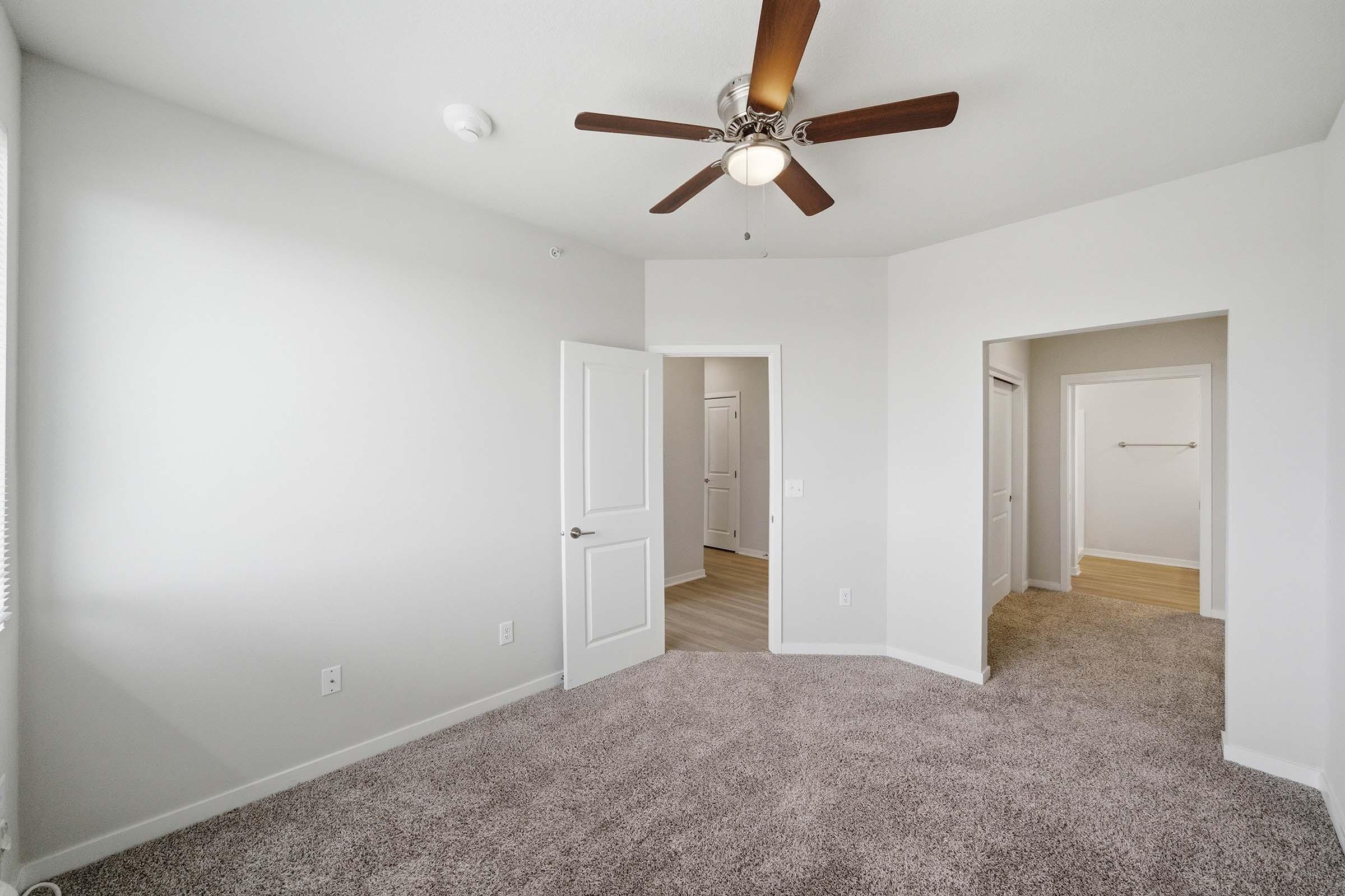 Bright and spacious room featuring light gray walls, plush carpeting, and a ceiling fan. The room includes a closed door leading to another area, with an adjacent doorway that opens to a hallway. Natural light streams in through a window, creating a welcoming atmosphere.