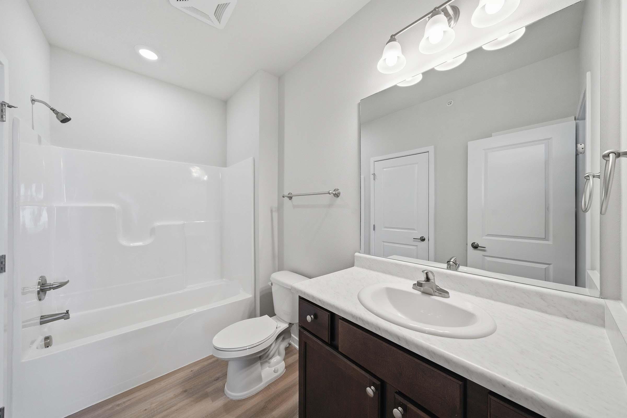 A modern bathroom featuring a white shower-tub combo, a single sink vanity with a marble countertop, a large mirror, and white cabinetry. The flooring is light wood, and the walls are painted in a neutral tone. Chrome fixtures add a contemporary touch to the space.