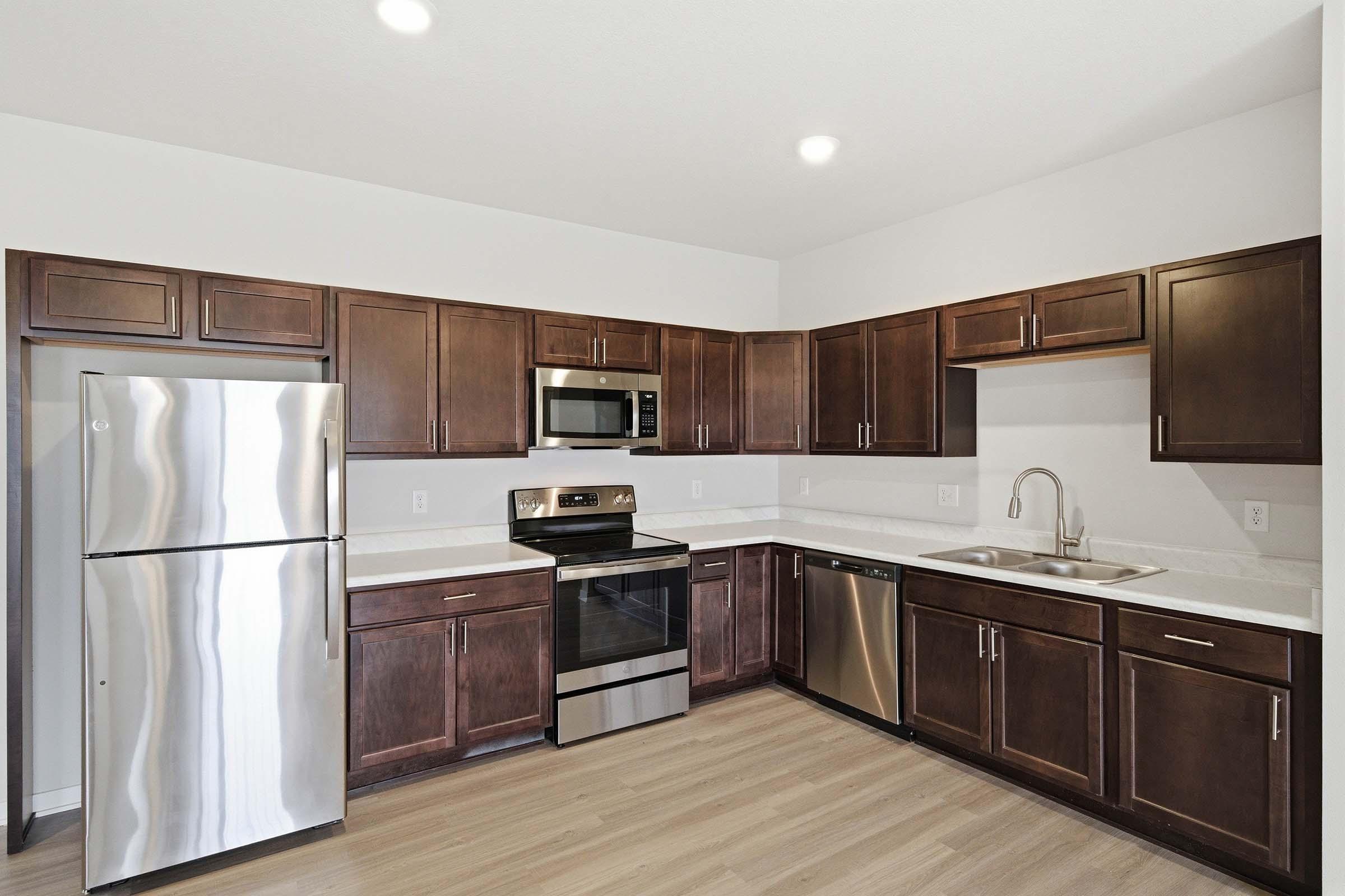 Modern kitchen featuring dark wood cabinets, stainless steel appliances including a refrigerator, microwave, oven, and dishwasher. The countertop is light-colored, contrasting with the dark cabinets, and the flooring is a light wood finish. Bright overhead lights illuminate the space.