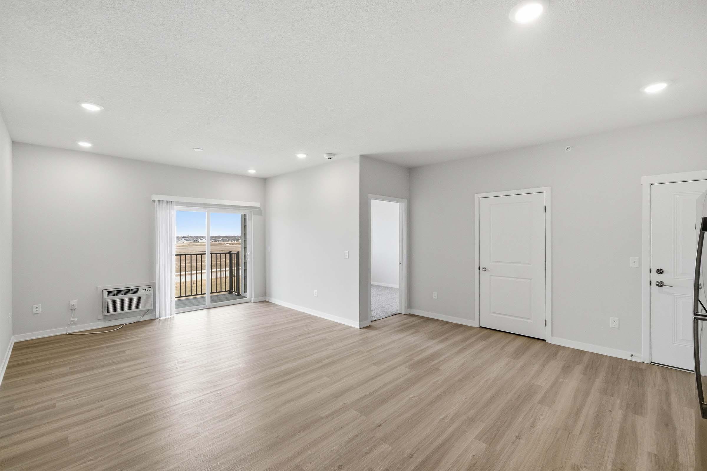 Spacious, well-lit living room featuring light-colored walls and laminate flooring. Includes a sliding glass door leading to a balcony with scenic views, an air conditioning unit, and an entrance to a neighboring room. Fresh and modern design with ample natural light.