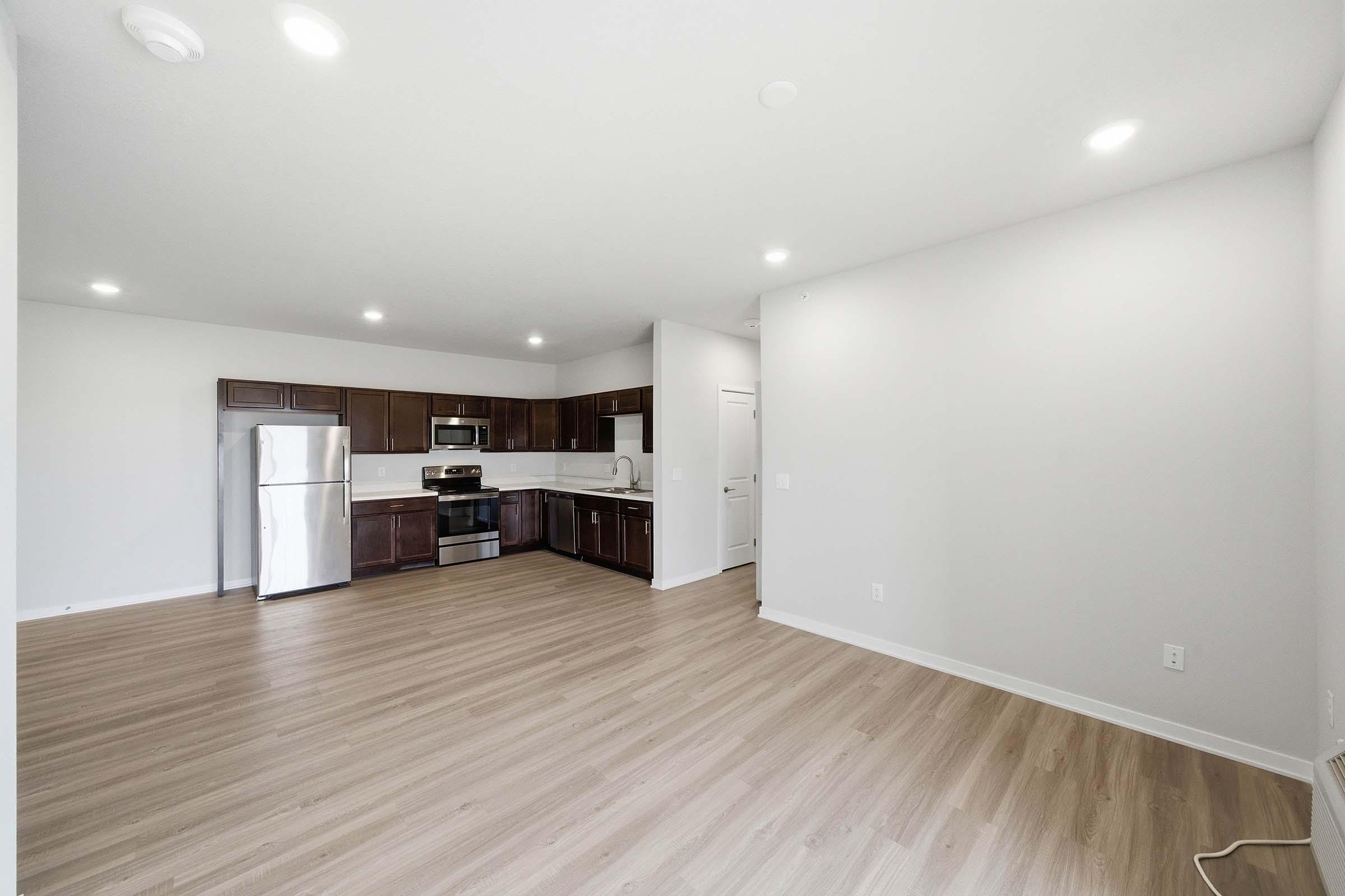 Modern kitchen and living area featuring dark wooden cabinets, stainless steel appliances including a refrigerator and oven, and a spacious open layout with light-colored walls and flooring. Bright overhead lighting enhances the airy feel of the space.