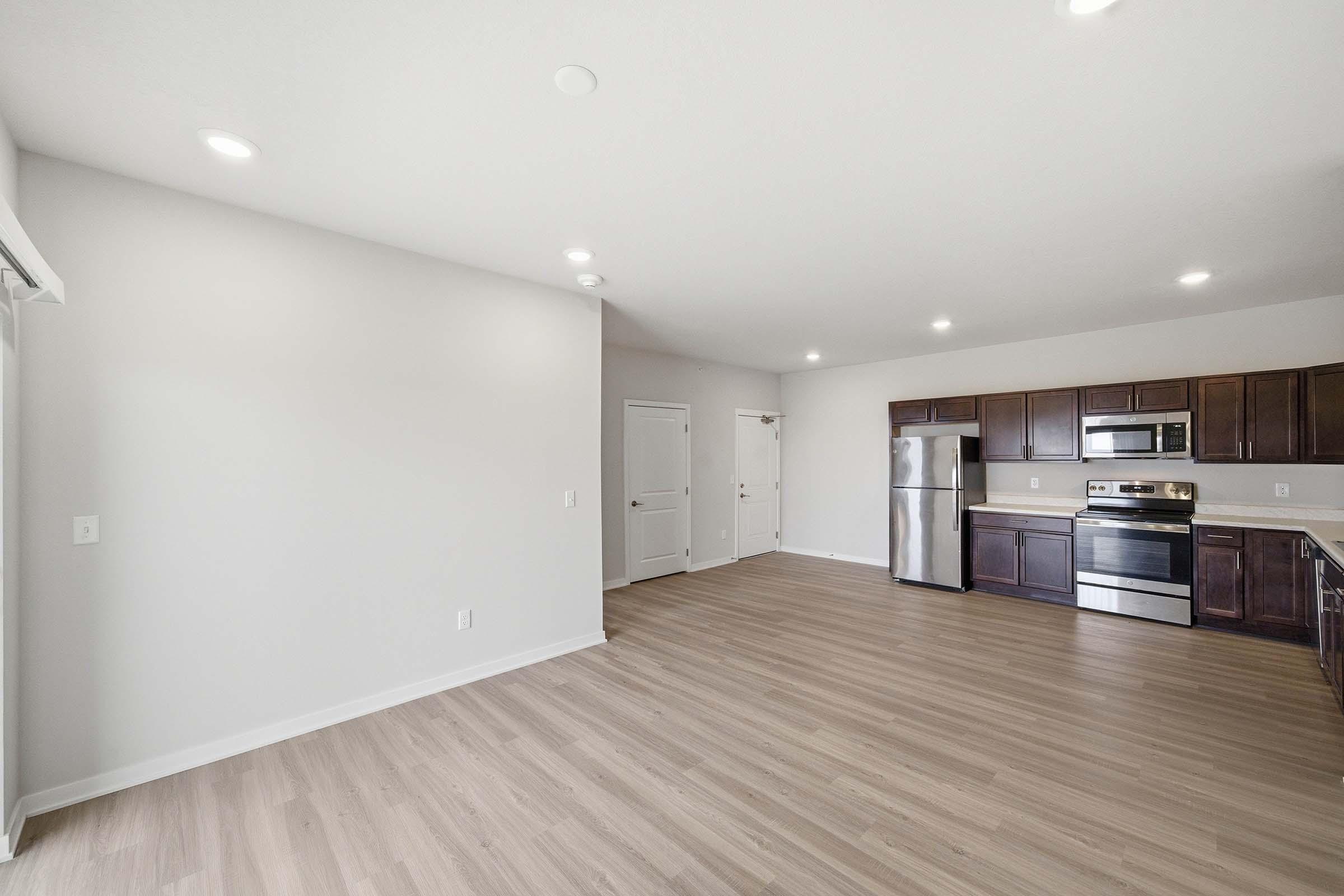 A spacious, well-lit interior of an open-concept kitchen and living area. The kitchen features dark wooden cabinets, stainless steel appliances, and a counter adjoining the living space. The floors are light-colored wood, and the walls are painted in a neutral tone. Windows allow natural light to fill the room.