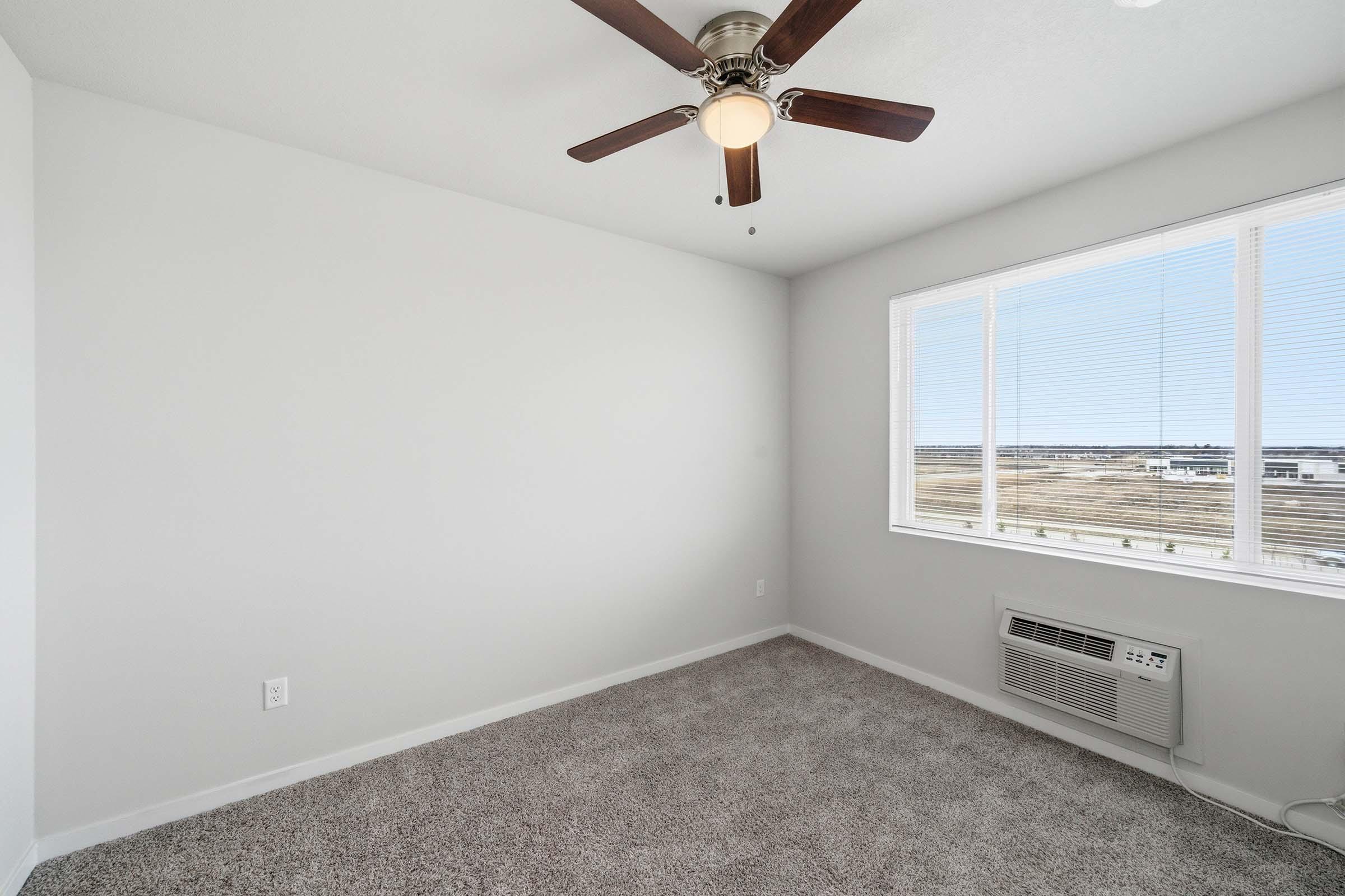A bright, empty room with light gray walls and a ceiling fan. The space features a large window with blinds, offering a view of the outside landscape. The floor is covered with plush, light-colored carpet, and there's an air conditioning unit mounted beneath the window.